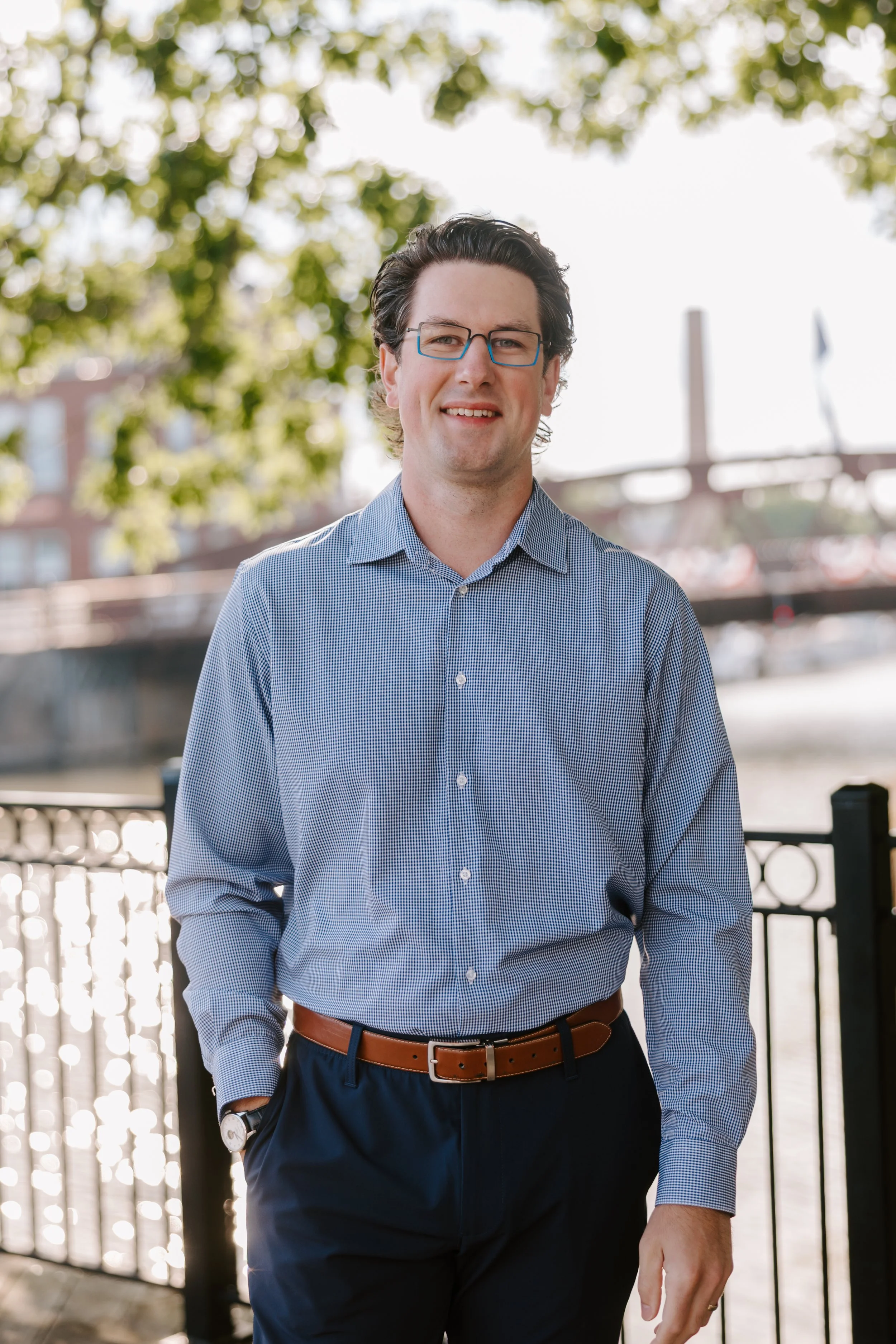 A man with glasses, wearing a blue checkered dress shirt and navy pants, walking outdoors near a railing with trees and a bridge in the background.