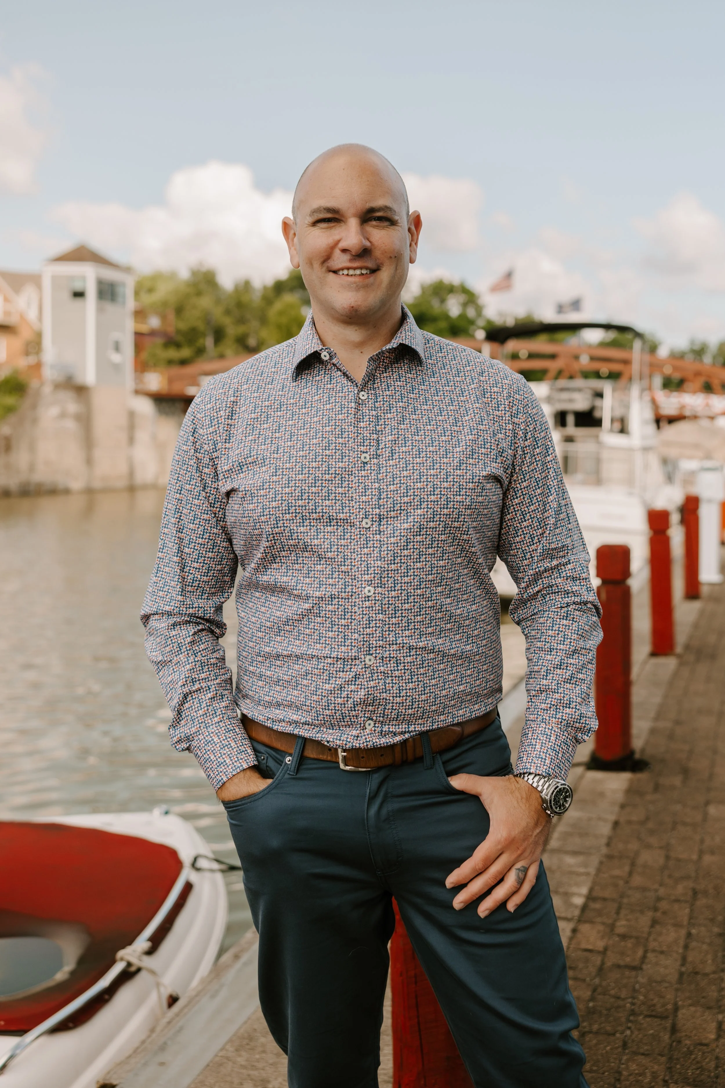 A man wearing a patterned button-up shirt and dark pants standing on a waterfront dock with boats in the background, smiling at the camera.