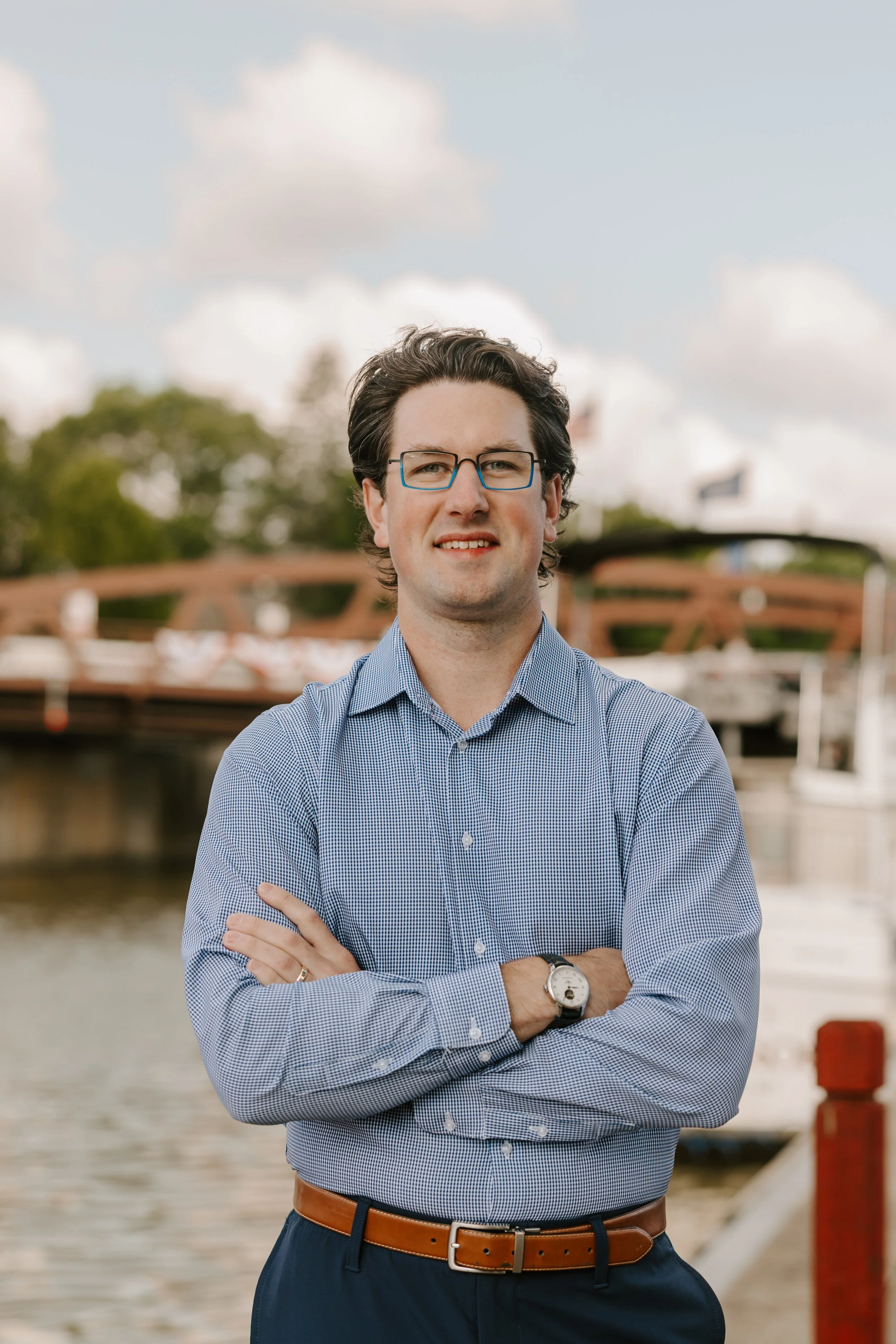 A man with brown hair, glasses, wearing a blue checkered shirt and brown belt, standing with arms crossed in front of a canal with a bridge and boats in the background, under a partly cloudy sky.