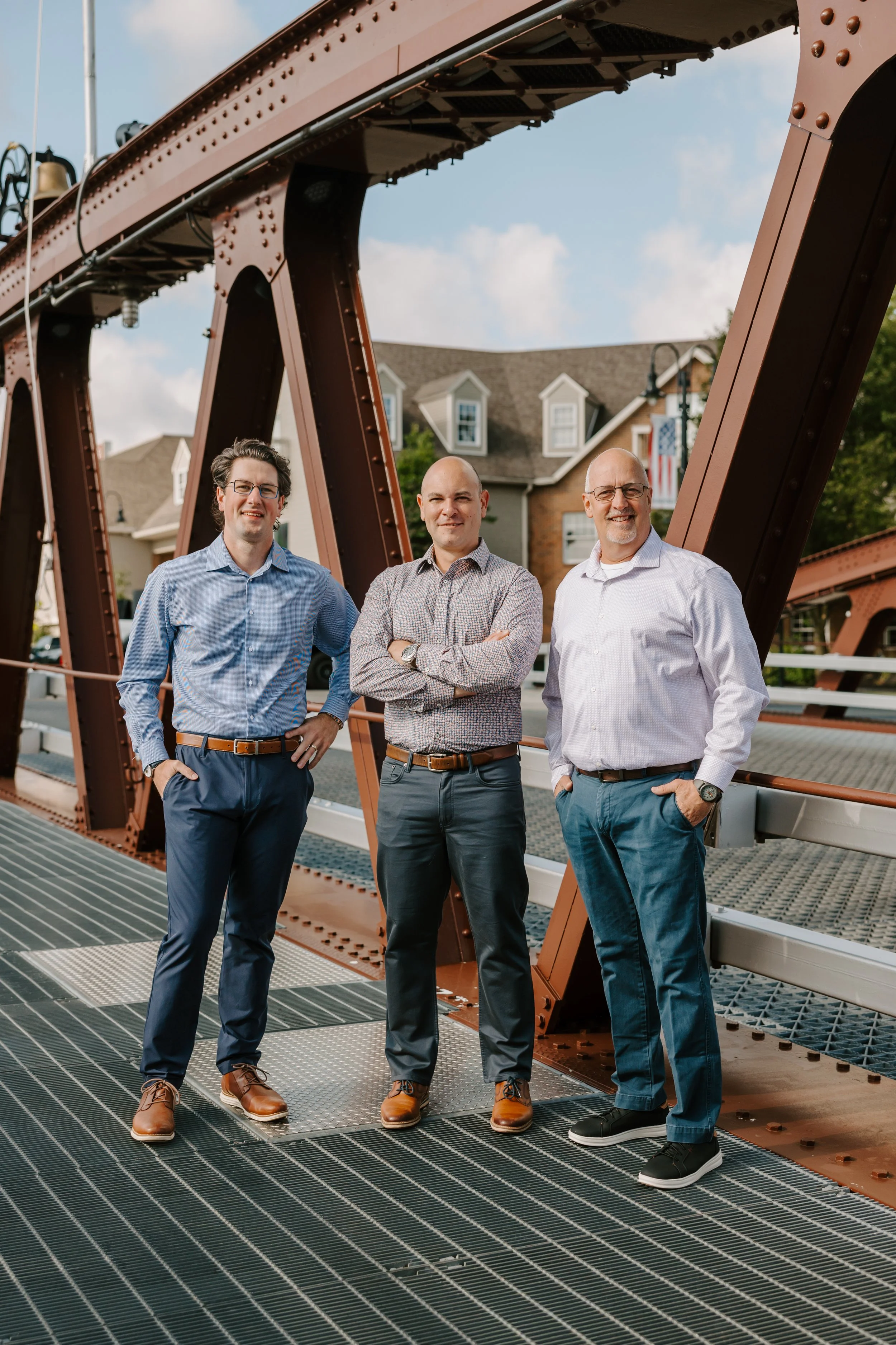 Three men standing on a metal bridge with a background of residential houses and a blue sky.