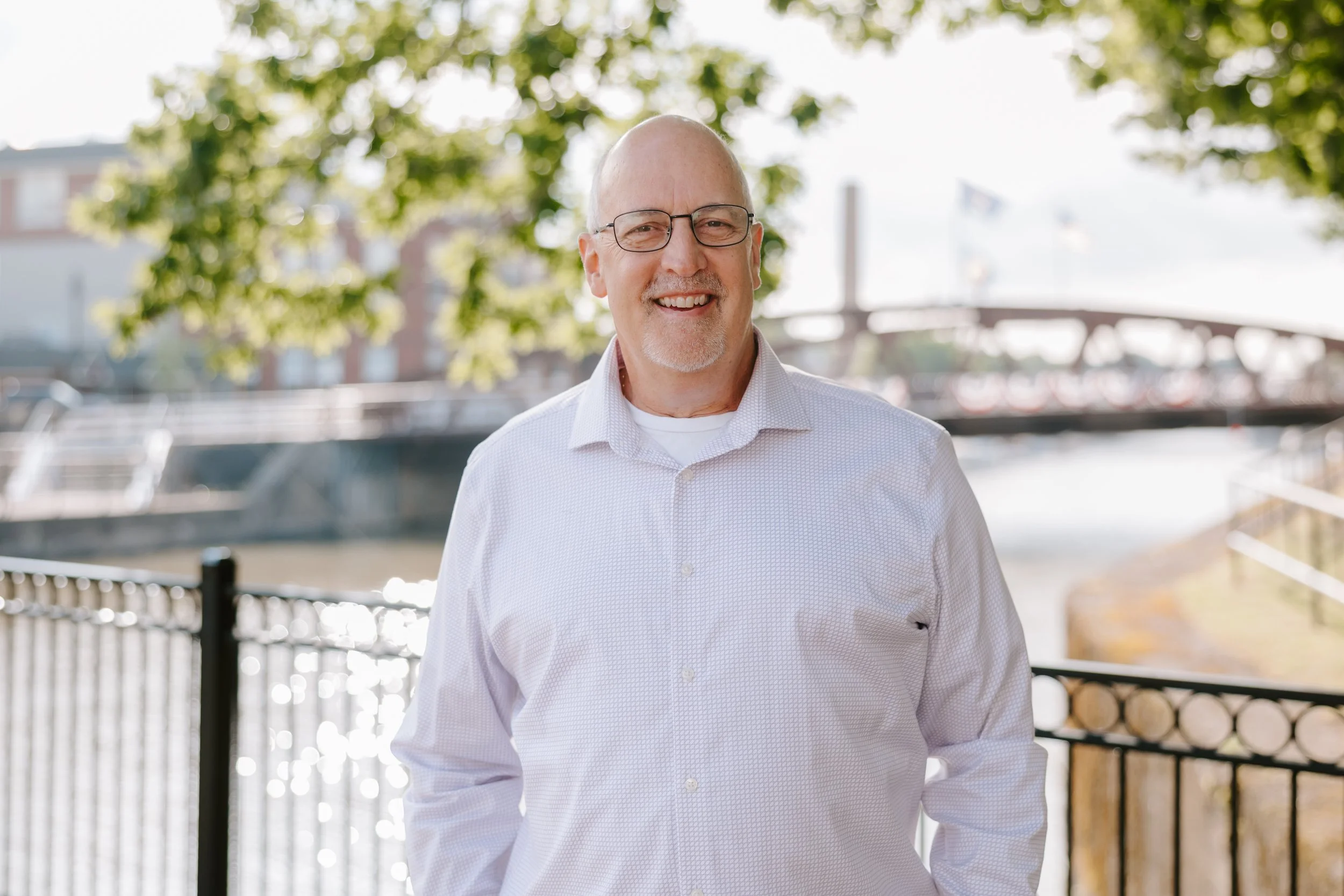 A smiling bald man with glasses in a white shirt outdoors, with trees and a fence in the background.
