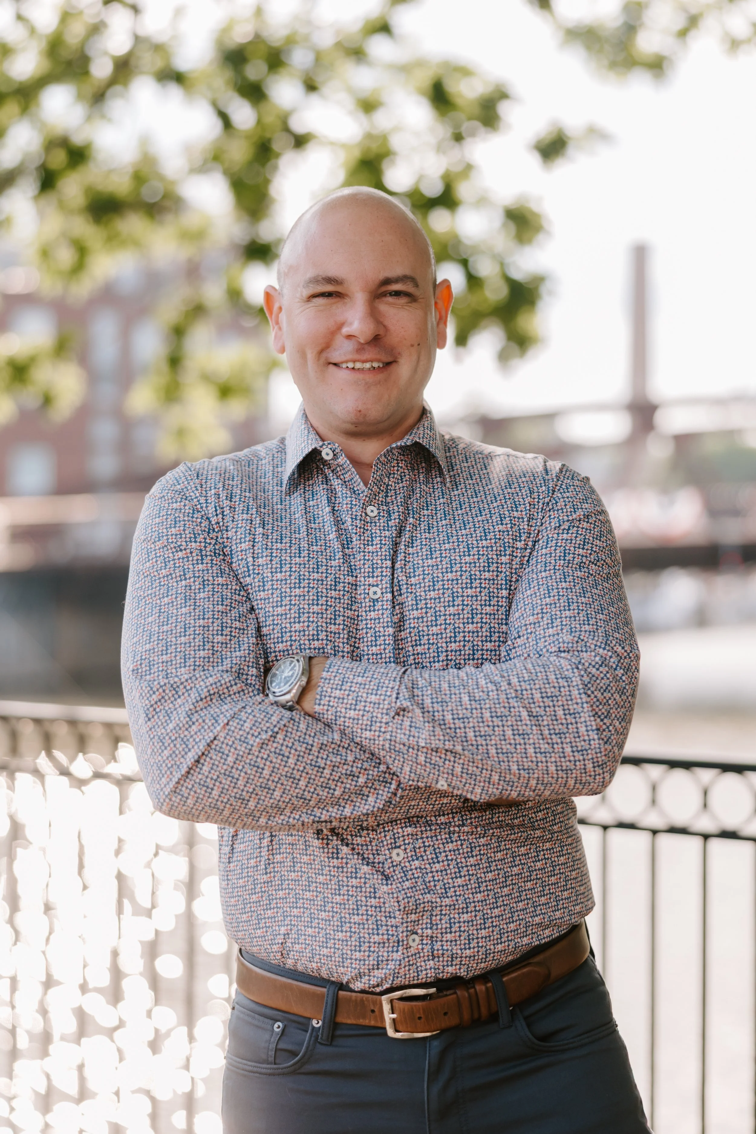 A smiling bald man in a patterned shirt with arms crossed, standing outdoors near a railing with green trees and a bright sky in the background.