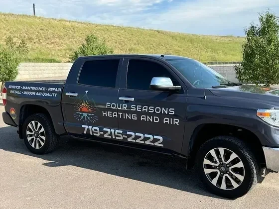 Black pickup truck with Four Seasons Heating and Air logo and contact number parked on street with grassy hillside and blue sky in background.