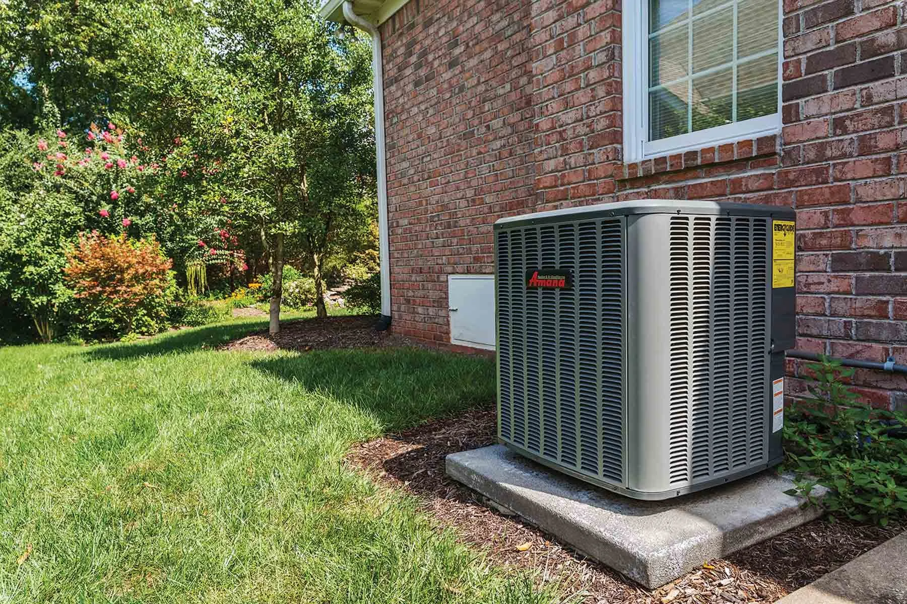 An outdoor scene showing a brick house with an air conditioning unit on a concrete pad, surrounded by green grass and trees with pink and green foliage.