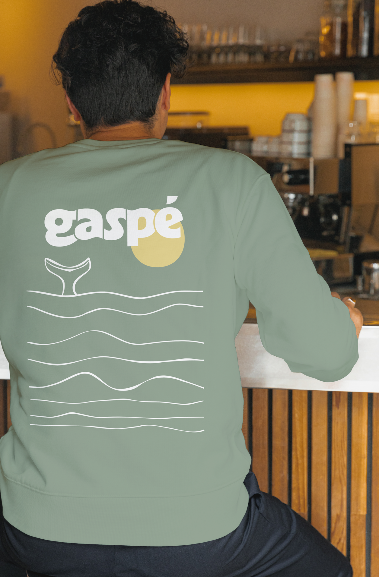 Une personne vue de dos, portant un sweat vert pâle avec le logo 'gaspé' et un dessin de vagues. La personne est assise au comptoir d'un café ou d'une bar, entourée d'étagères avec des tasses et des équipements en arrière-plan.