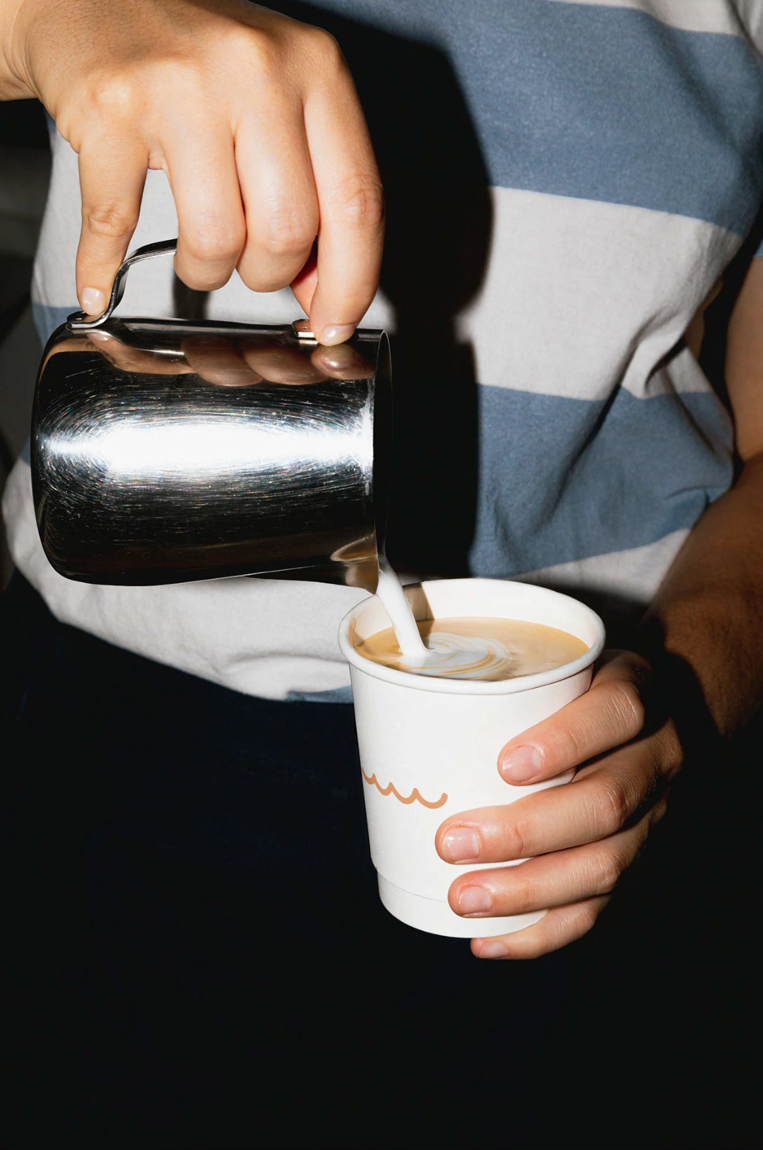 Une personne verse du lait dans une tasse de café, la personne tient une tasse en papier blanc avec un dessin de vagues orange, et porte une chemise à rayures bleues et blanches.