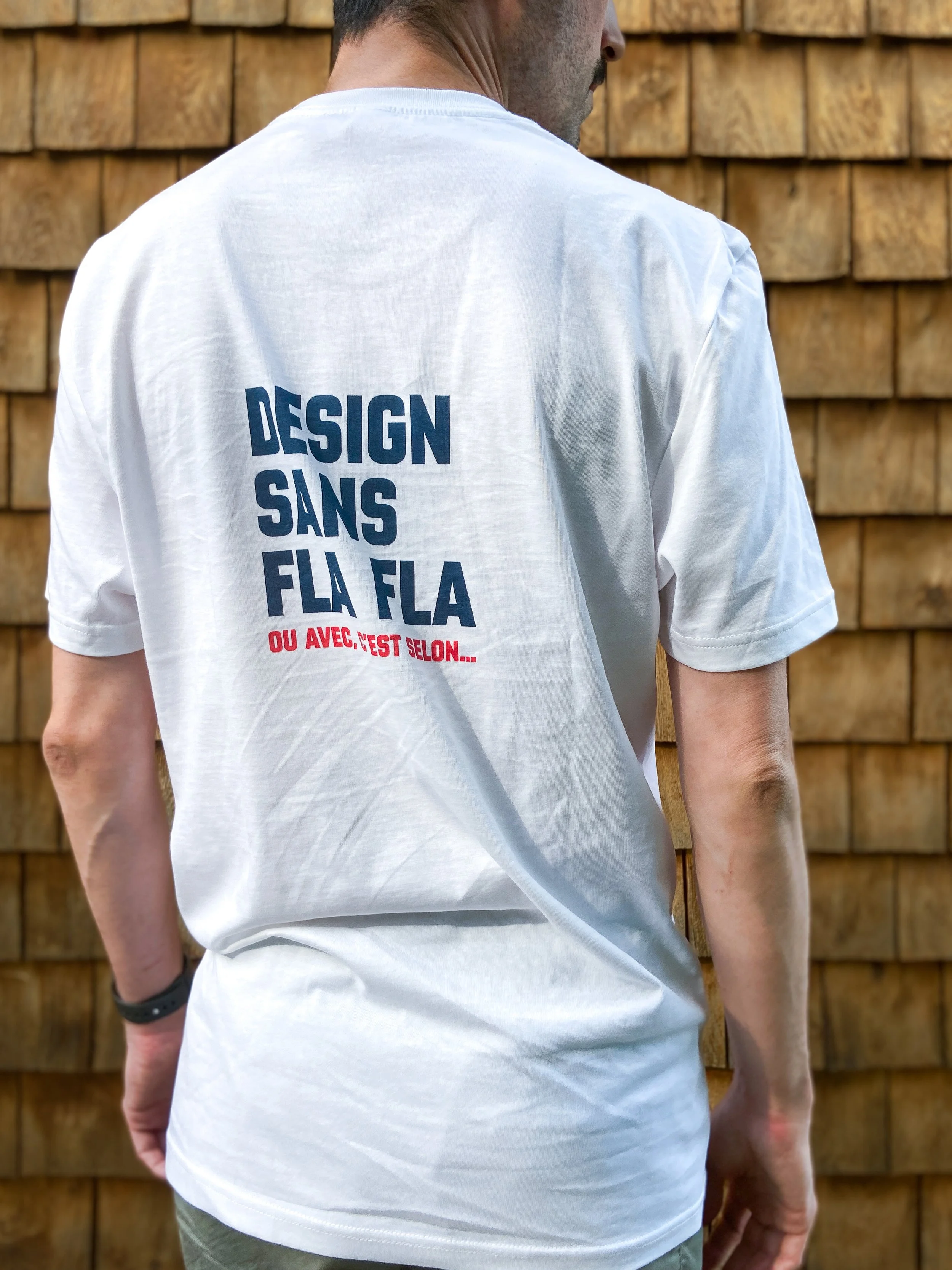 Personne portant un t-shirt blanc avec l'inscription «design sans fla fla, ou avec, c'est selon...» vue de dos contre un mur en bois.