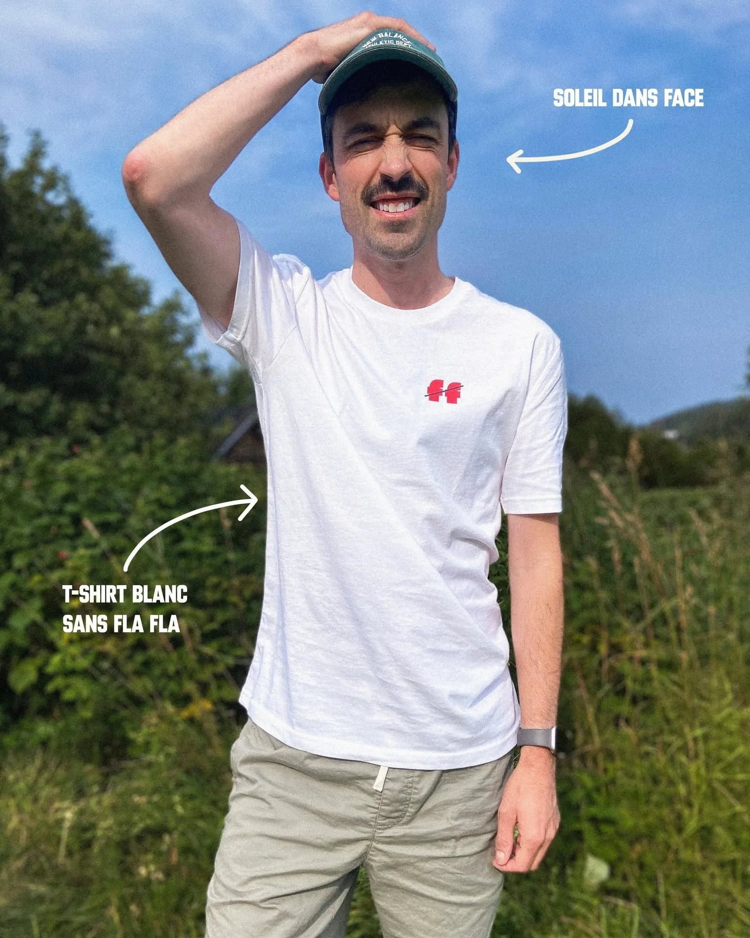 Un homme portant un t-shirt blanc avec logo, des pantalons beige, et une montre, se tenant dans la nature avec un ciel bleu en arrière-plan. Il met la main sur la tête et sourit légèrement.