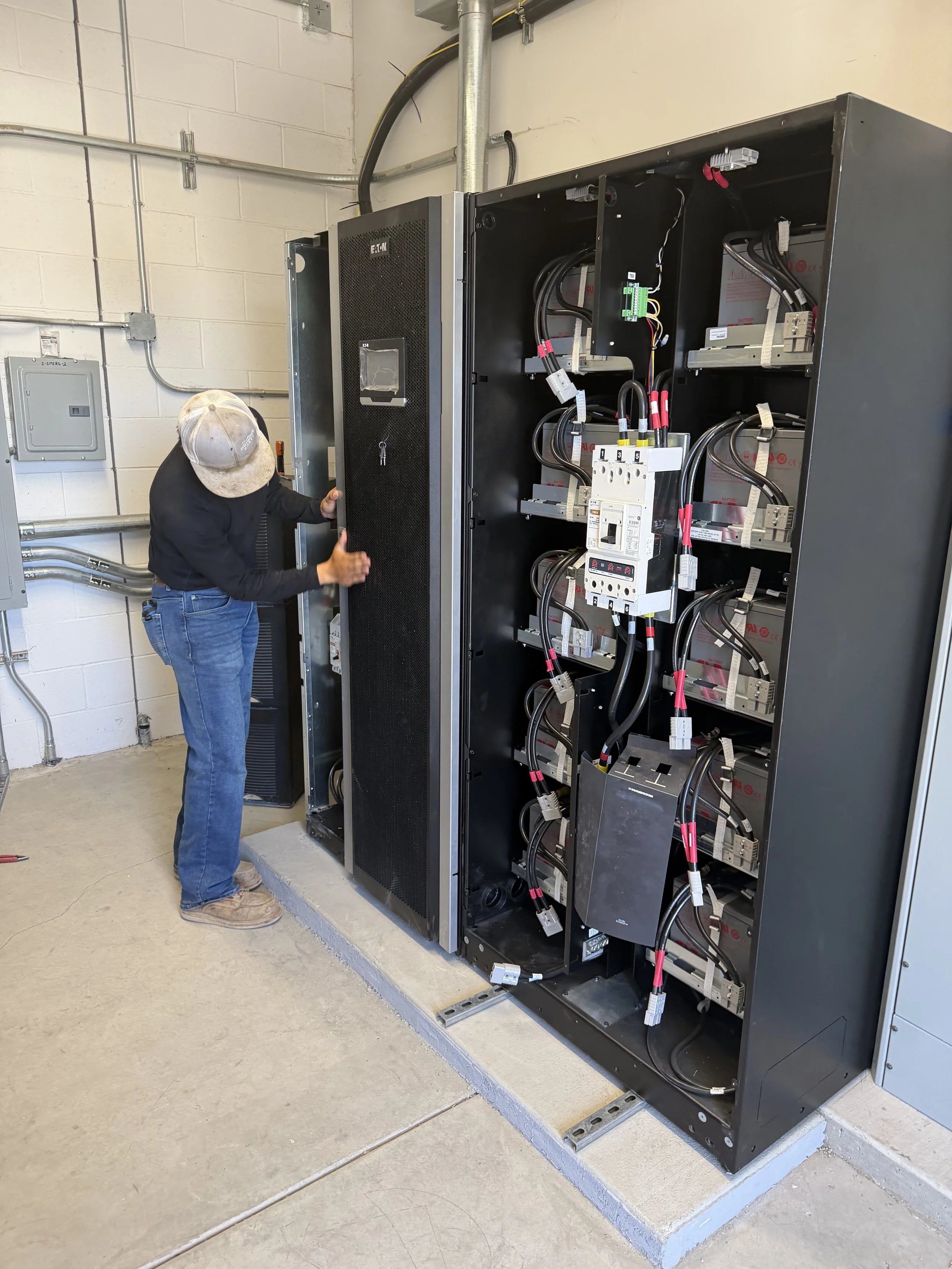 A technician working on an electrical panel with open doors, showing internal wiring and components, in an industrial or utility room with exposed conduit and a concrete floor.