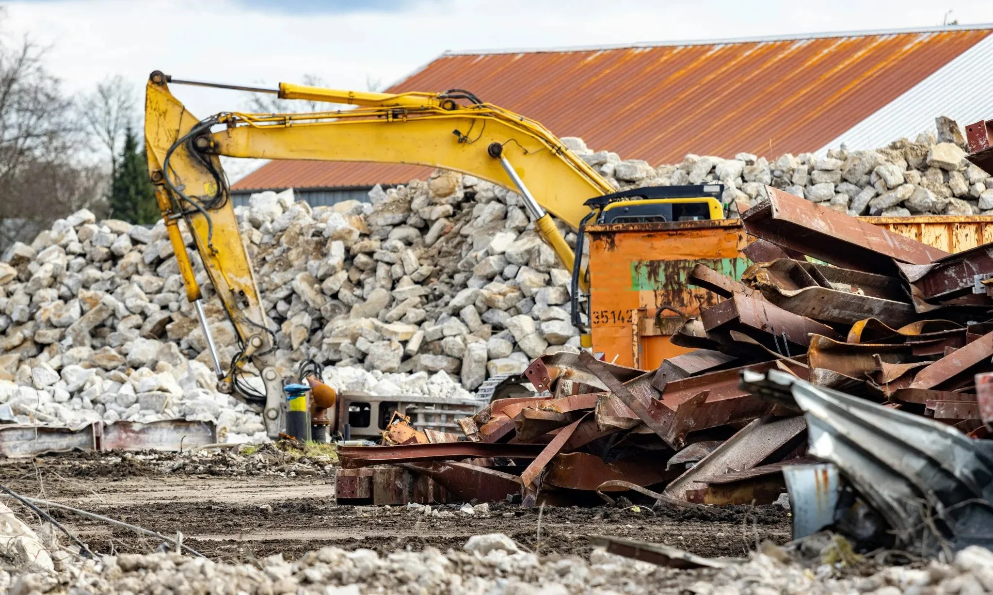 A construction site with a yellow excavator lifting large rocks, surrounded by piles of rubble and metal debris, near a building with a rust-colored roof.