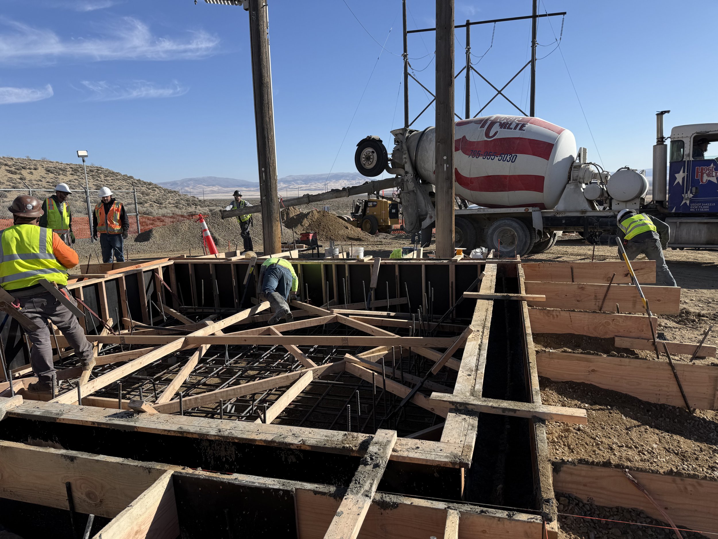 Construction workers in high-visibility vests and hard hats working on a framework for a concrete foundation on a construction site, with a cement mixer truck in the background under a clear blue sky