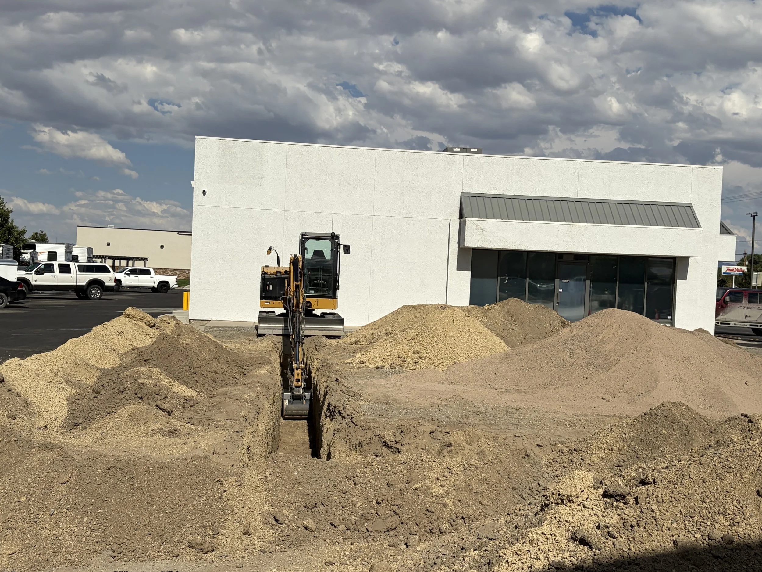 Construction site with dirt piles and a small excavator, adjacent to a white commercial building with parking lot and vehicles, under a partly cloudy sky.