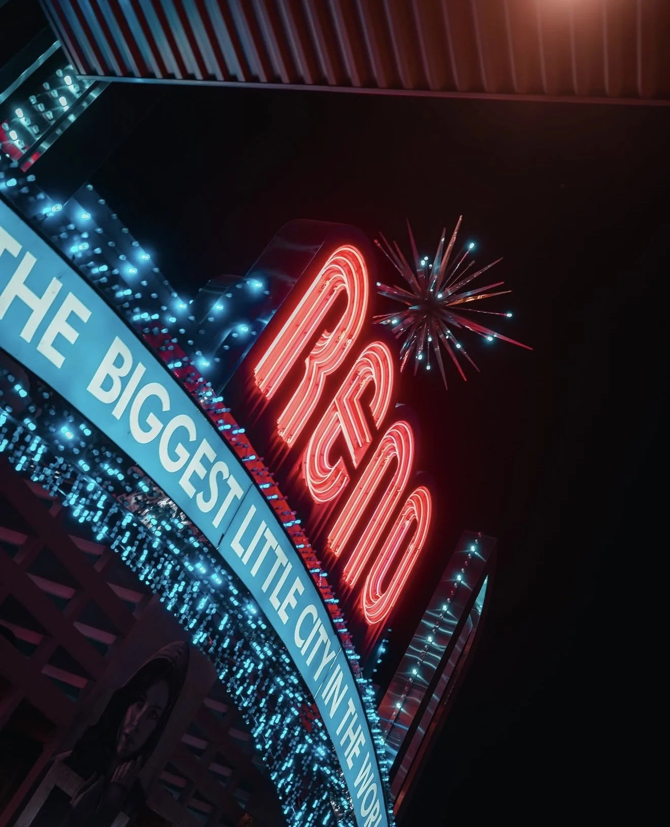 The image shows a brightly lit neon sign that reads 'RICO' in red letters, with fireworks in the background and a scrolling marquee at the bottom that says 'THE BIGGEST LIT...'. The scene appears to be at night, with colorful lights and decorations.