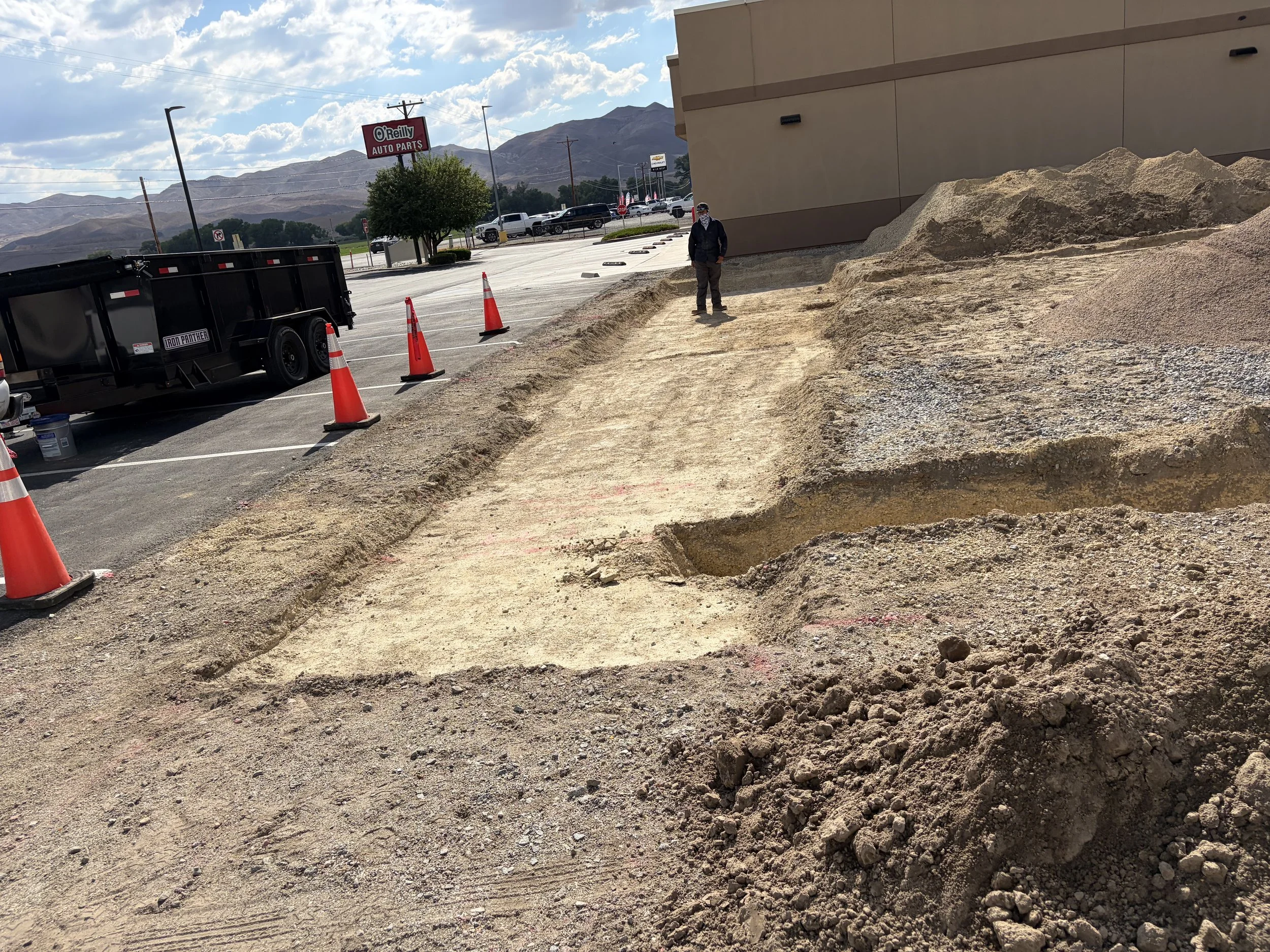 A sidewalk under construction with dirt piles, traffic cones, and a person standing nearby, next to a building and parking lot. Mountains and a partly cloudy sky in the background.