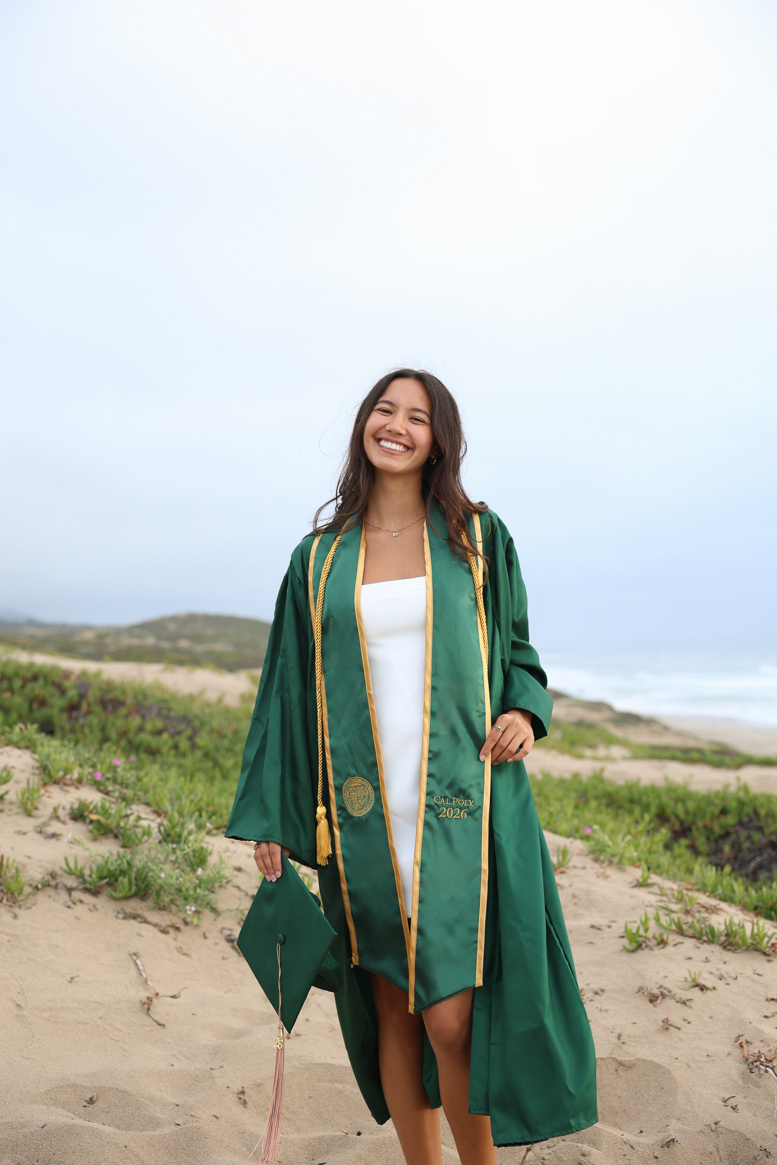 A young woman in a green graduation gown and cap standing on a sandy beach with green plants, holding her cap in her right hand and smiling at the camera.