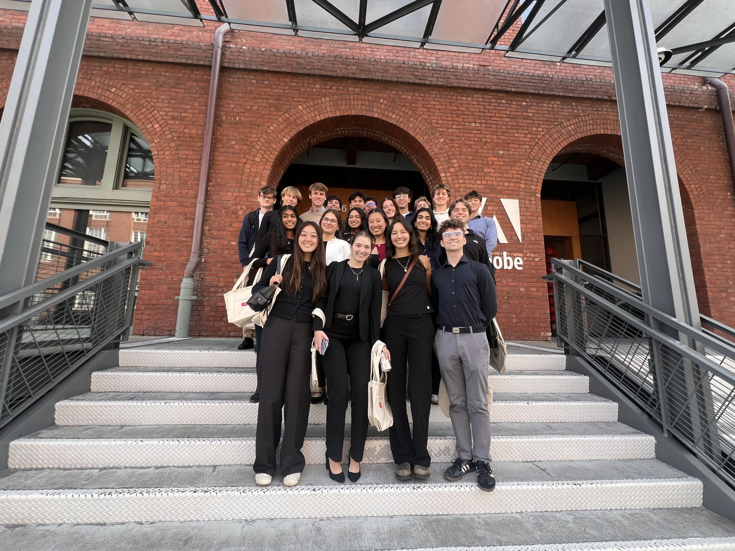 Group of diverse young adults standing on a metal staircase in front of a red brick building, smiling and posing for a group photo.