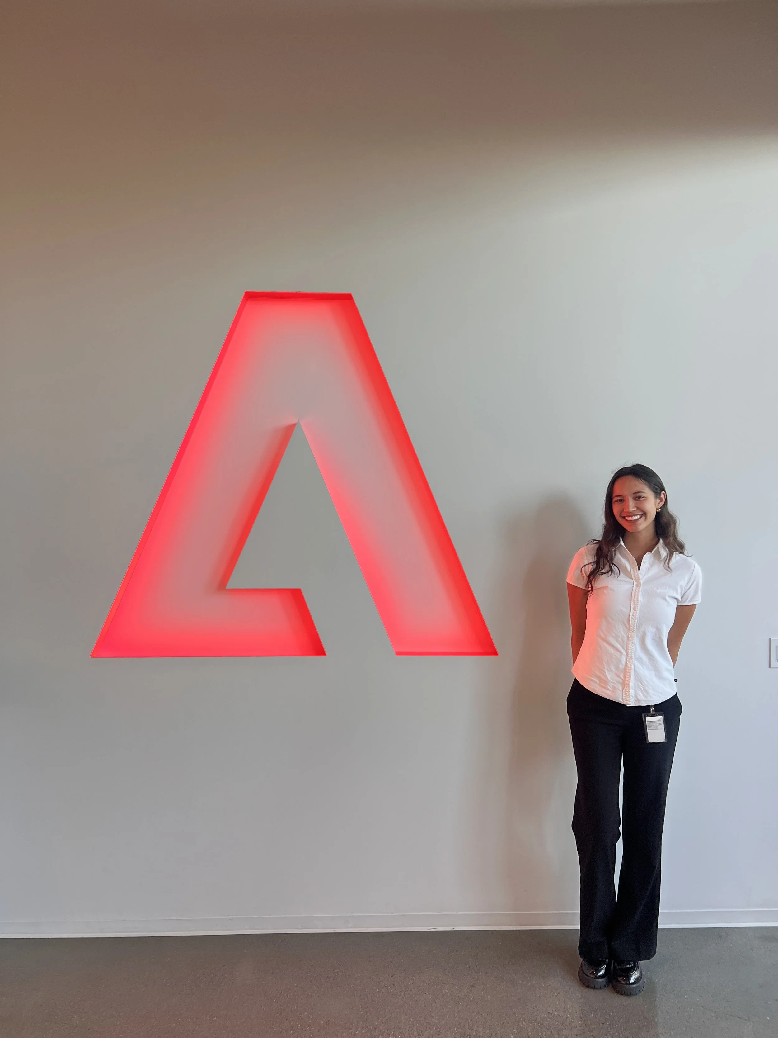 A young woman with long dark hair smiling and wearing a white short-sleeve shirt and black pants, standing next to a large red illuminated letter 'A' on a plain wall.