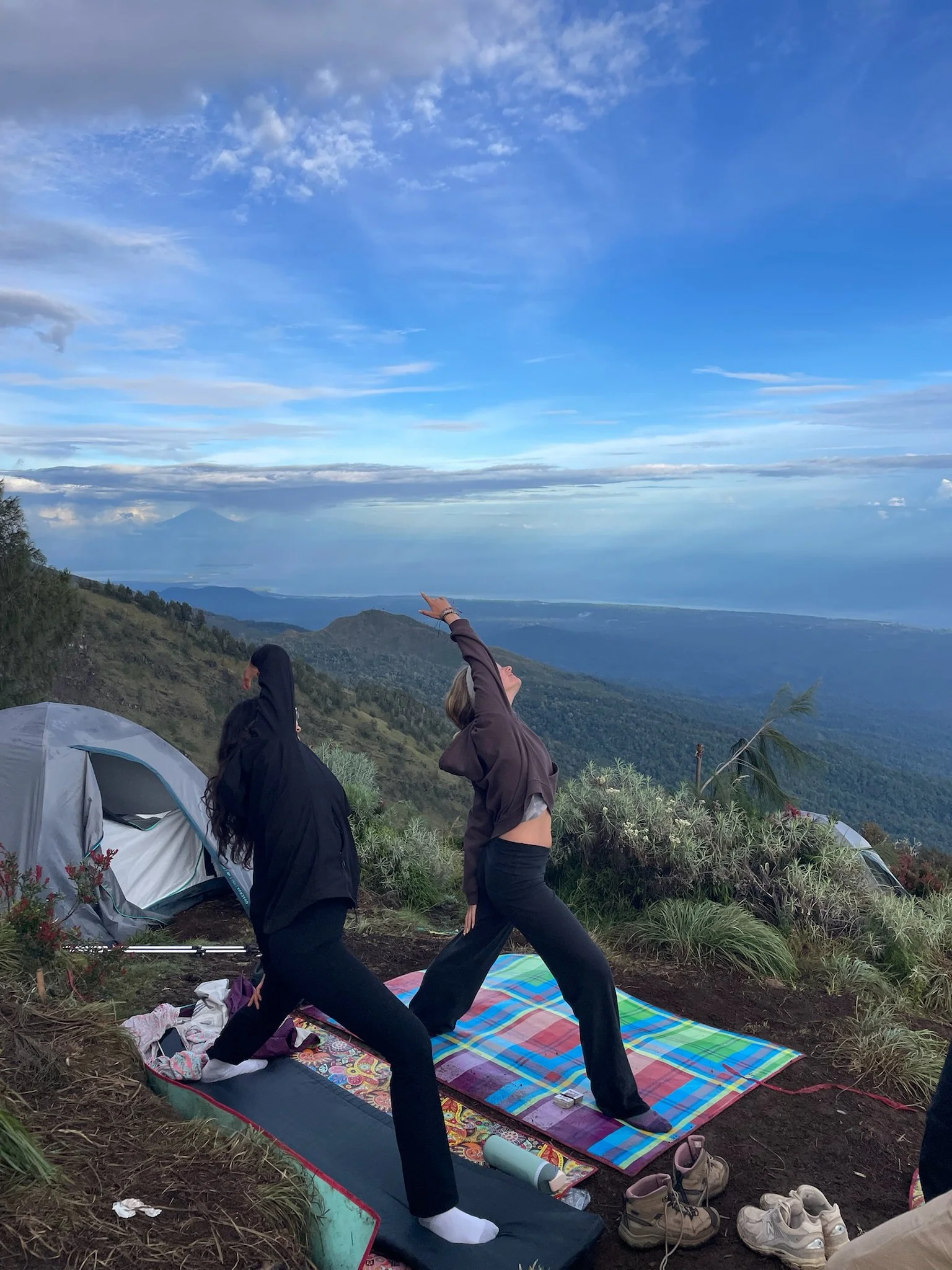 Two people practicing yoga outdoors on a colorful mat on a grassy hillside with tents and mountains in the background under a partly cloudy sky.