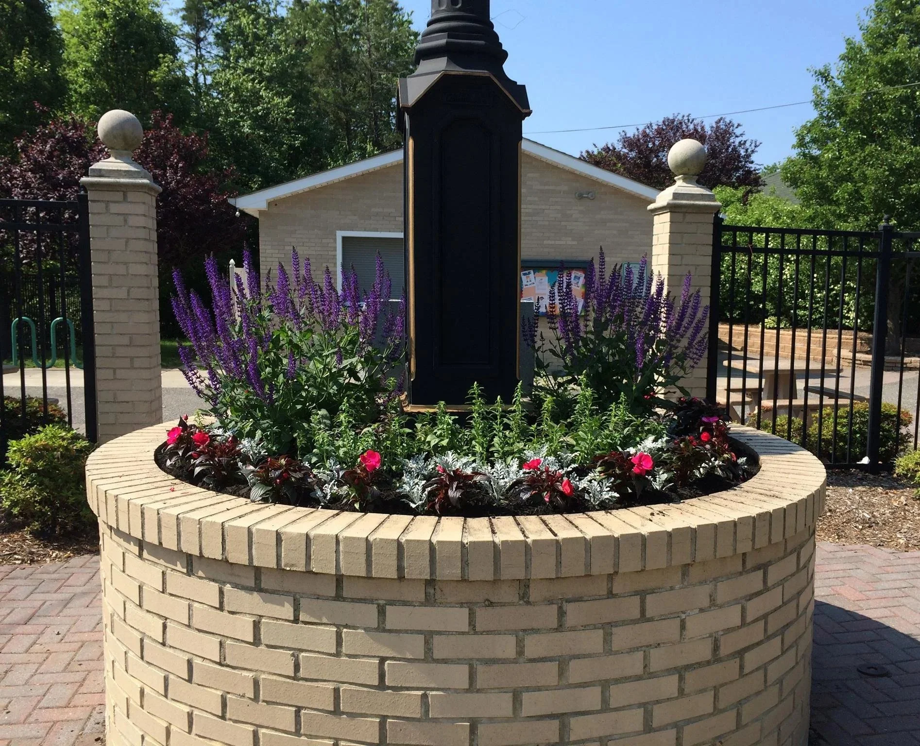 A decorative brick well with purple flowers surrounding it, fenced with black metal fence and situated in a garden area with trees and a small building in the background, under a clear blue sky.