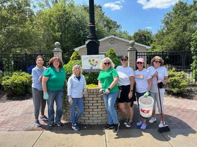 Group of seven women standing outdoors in front of a brick planter, with some holding cleaning tools, participating in a community cleanup event on a sunny day.