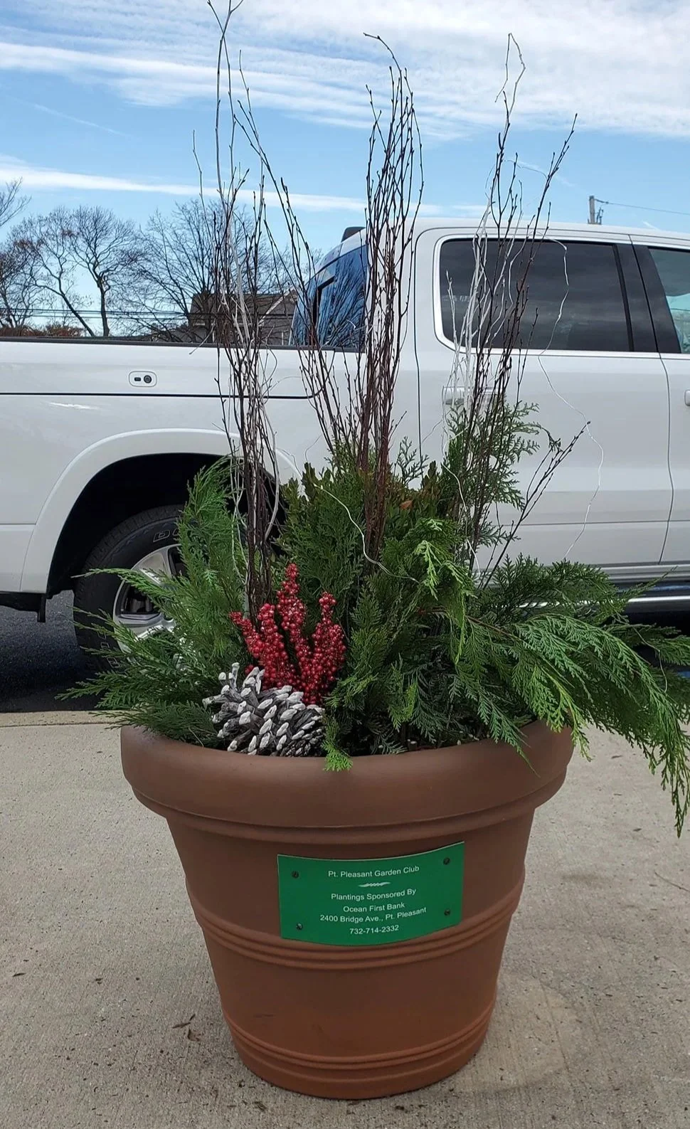Large terracotta planter with greenery and decorative elements, placed outside on pavement in front of a white pickup truck and a partly cloudy sky.
