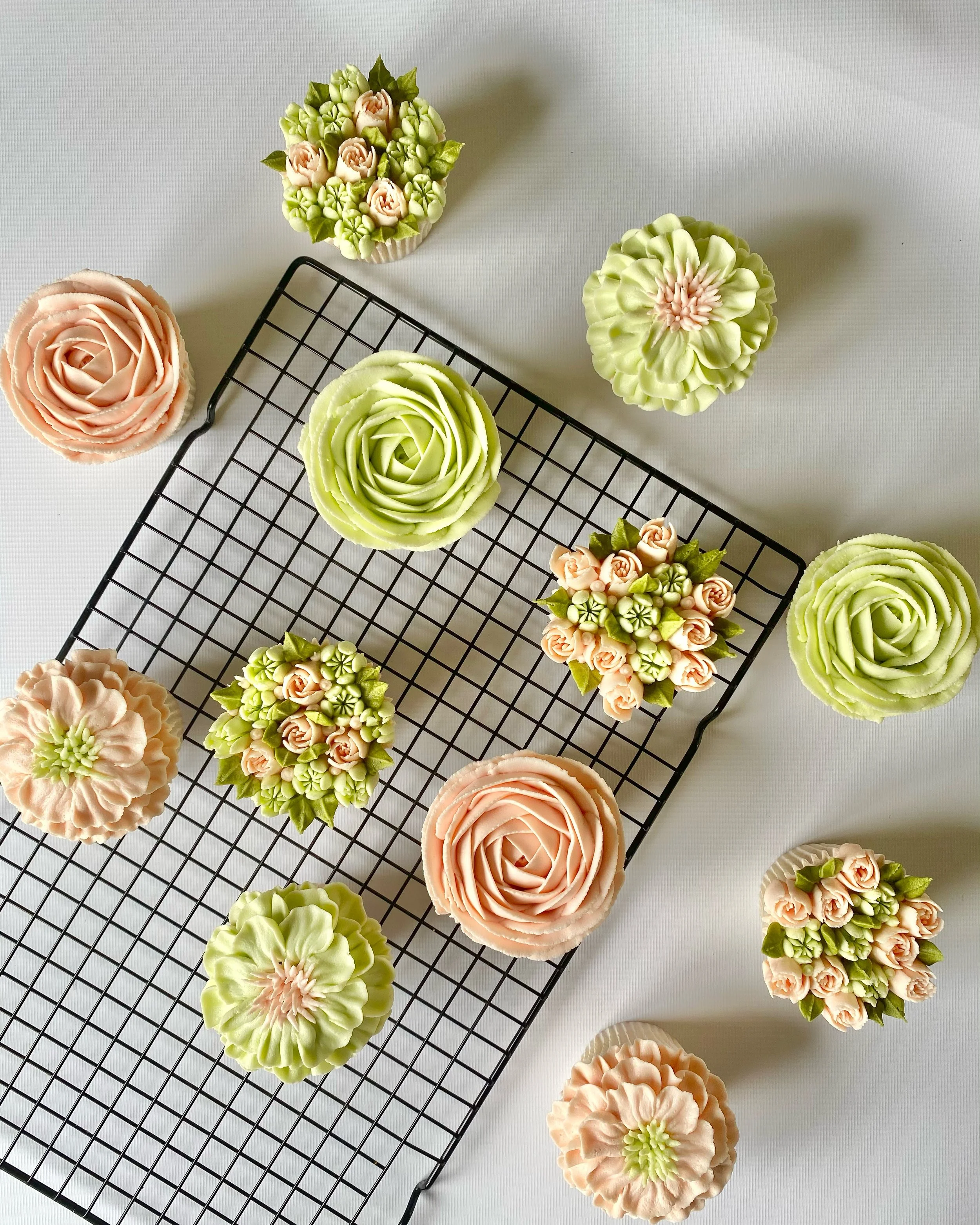 Assorted cupcakes decorated with floral and rose designs on a cooling rack, with more cupcakes and a white surface in the background.