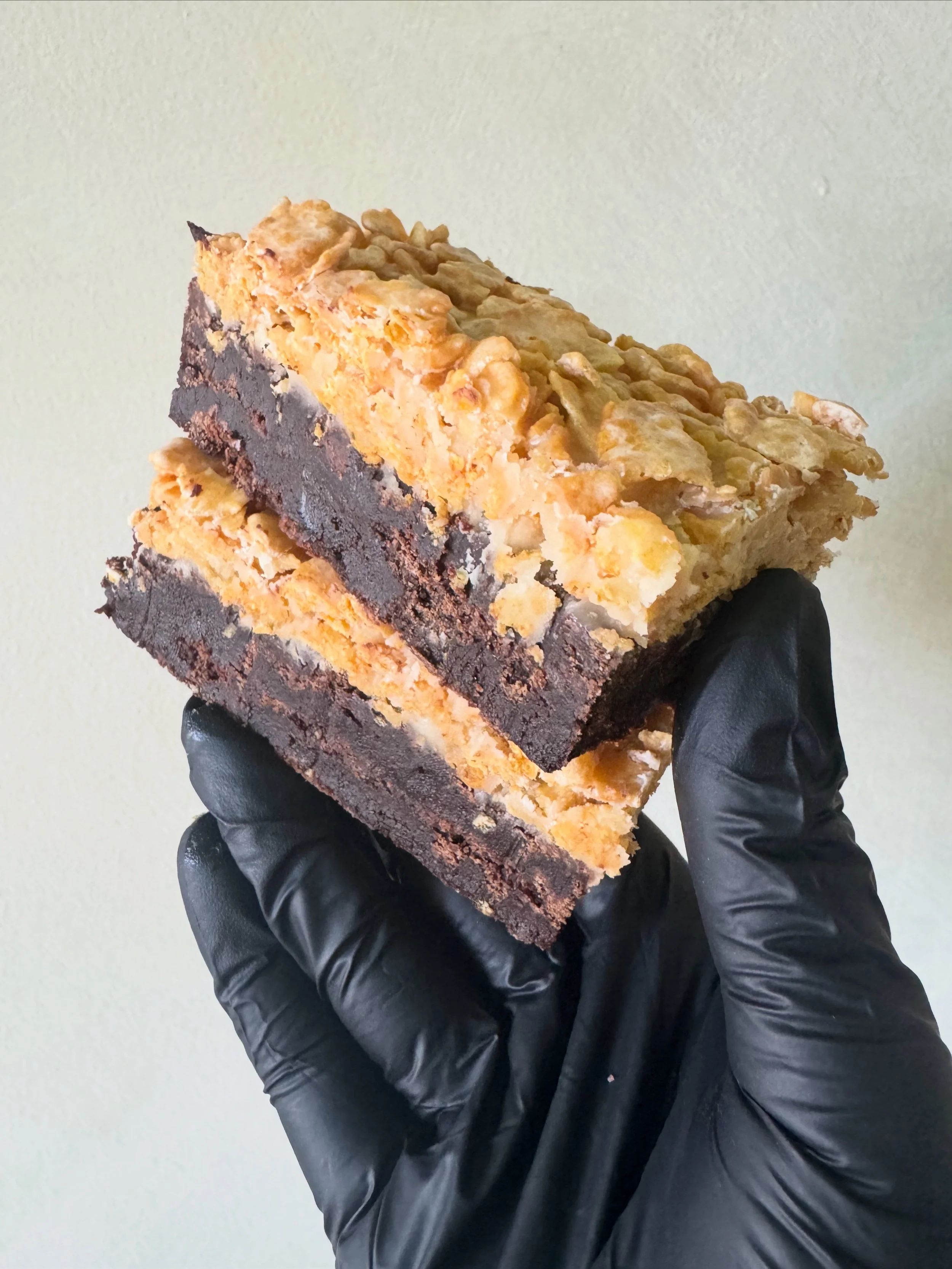 Close-up of a person holding a slice of layered dessert with a dark chocolate crust, caramel or butterscotch filling, and a topping of crumbled graham crackers or similar crumbs, against a plain light-colored background.