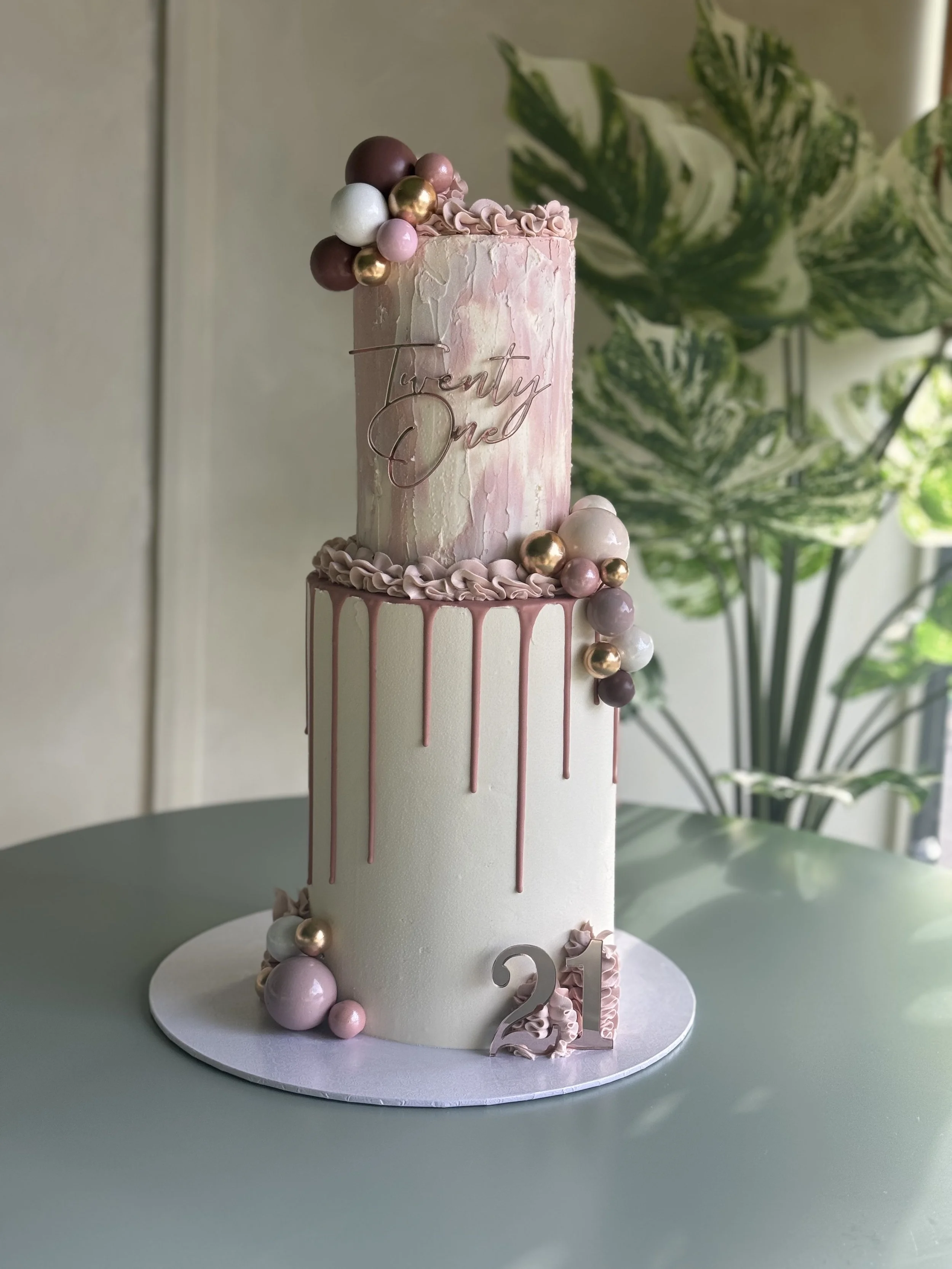 A tall, elegant birthday cake with pastel pink and white marbled icing, decorated with pink, gold, and white spherical candies, with a gold wire topper spelling 'twenty one,' and a pink drip around the top edge, set on a white circular base, in front of a leafy green plant.