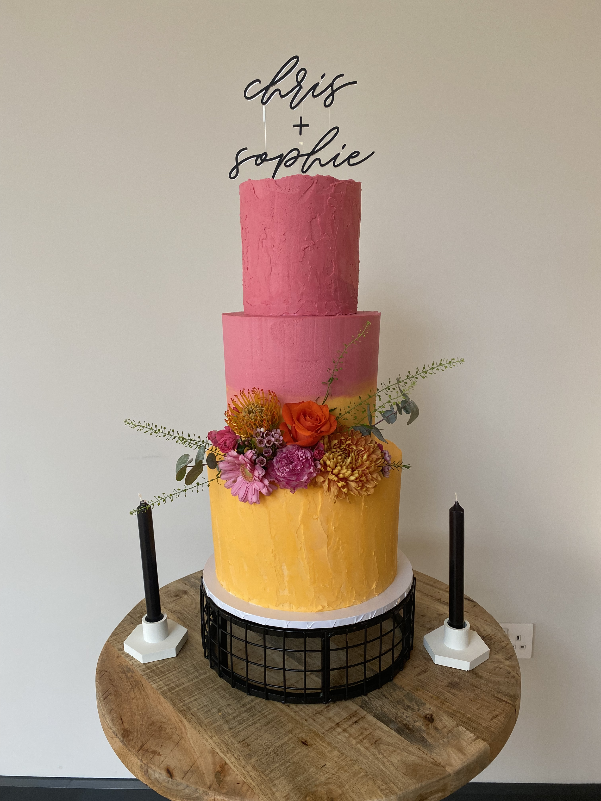 Three-tiered cake with a yellow bottom tier, pink middle tier, and pink top tier, decorated with fresh flowers, on a wooden table. Two black candles with white bases are on either side of the cake. A cake topper reads 'Chris + Sophie' in black cursive and print lettering.