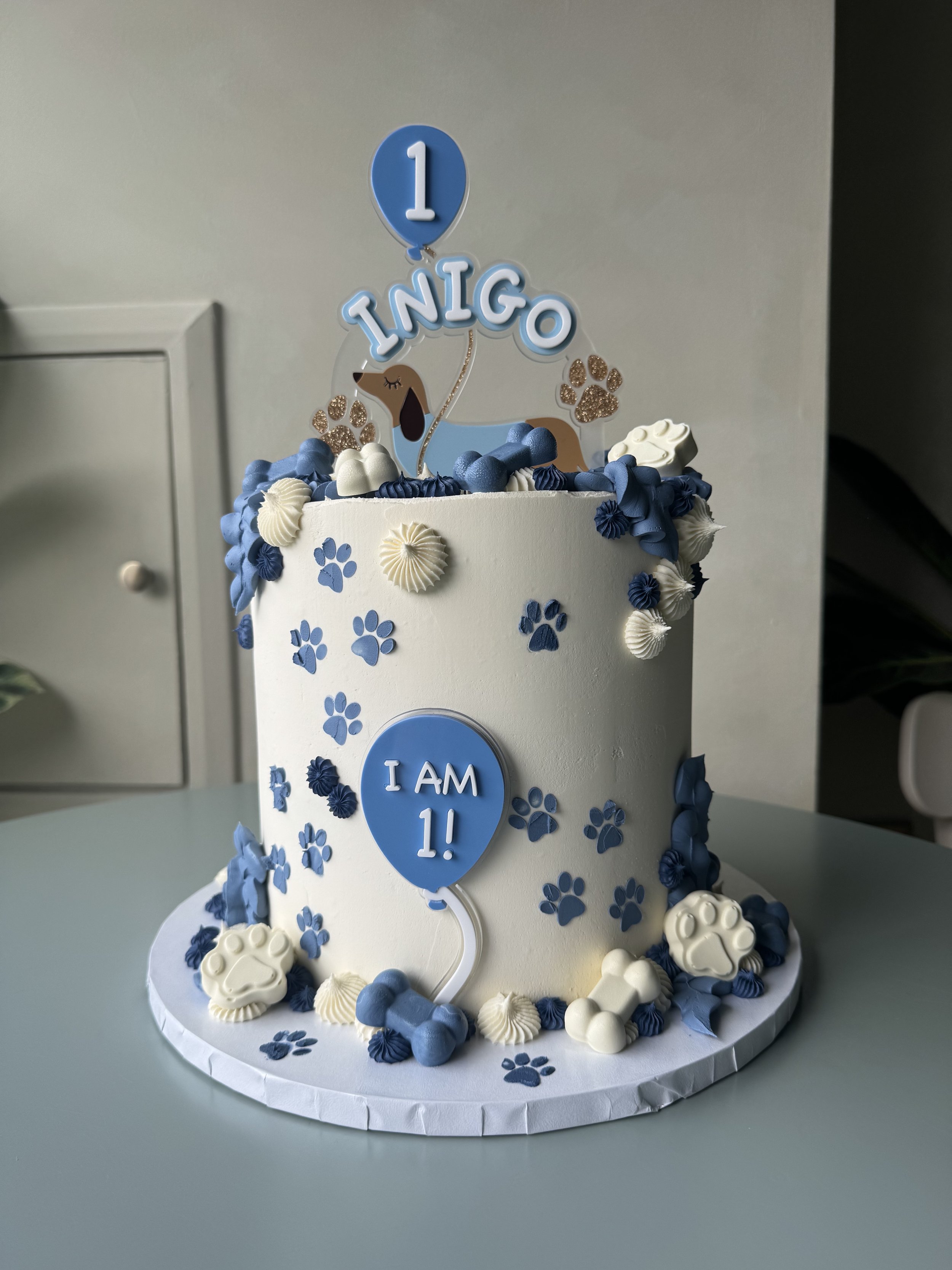 Celebrate a one-year-old's birthday with a white cake decorated with blue and white paw prints, shells, and a cake topper with a dog illustration, blue balloons, and the message 'I am 1!'.