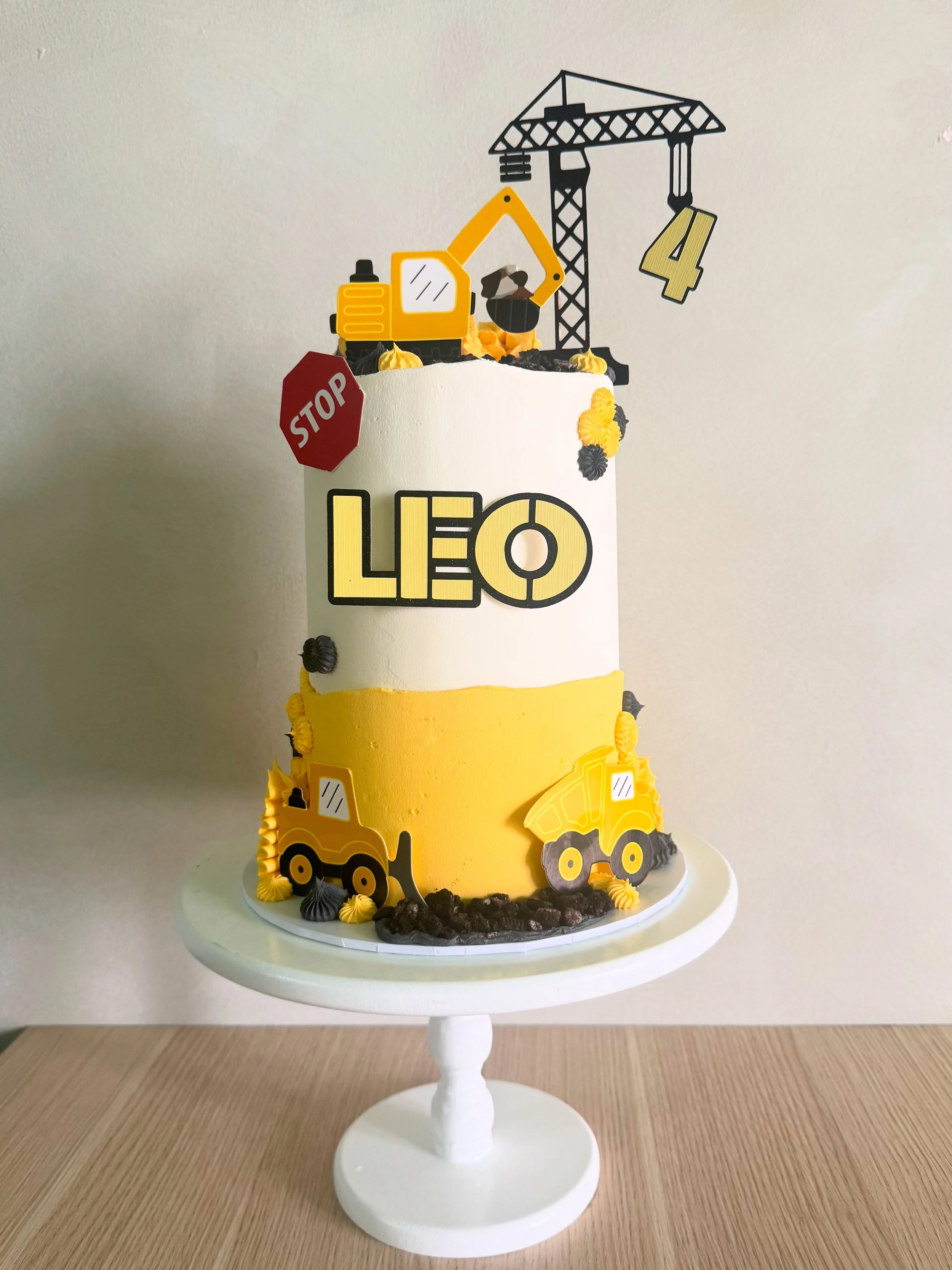 A construction-themed birthday cake with yellow and black construction vehicles, a stop sign, and construction equipment decorations on top, with a white and yellow color scheme.