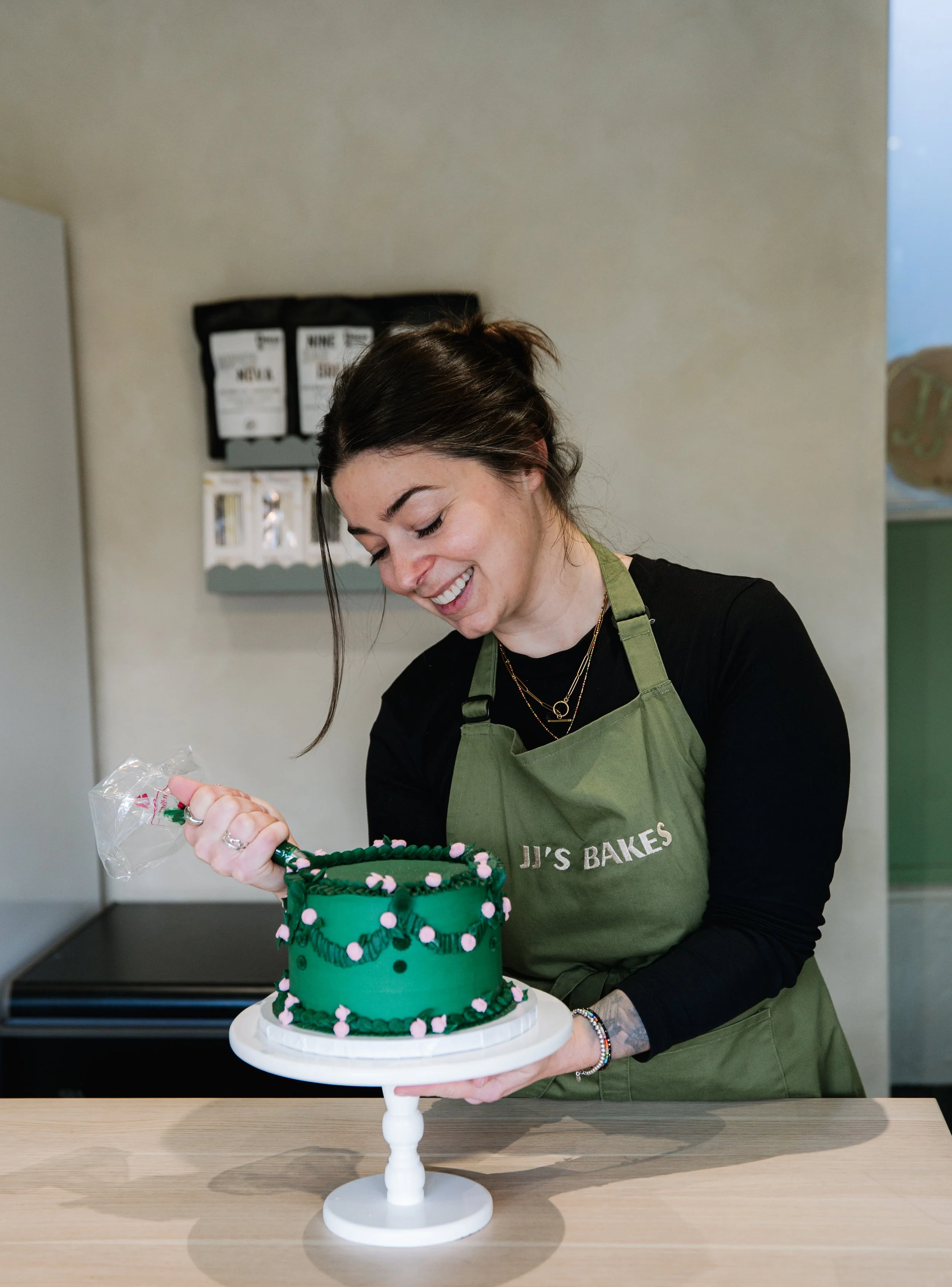 A woman is decorating a green cake with pink and black accents. She is smiling and wearing a green apron with 'JJ's Bakes' on it.