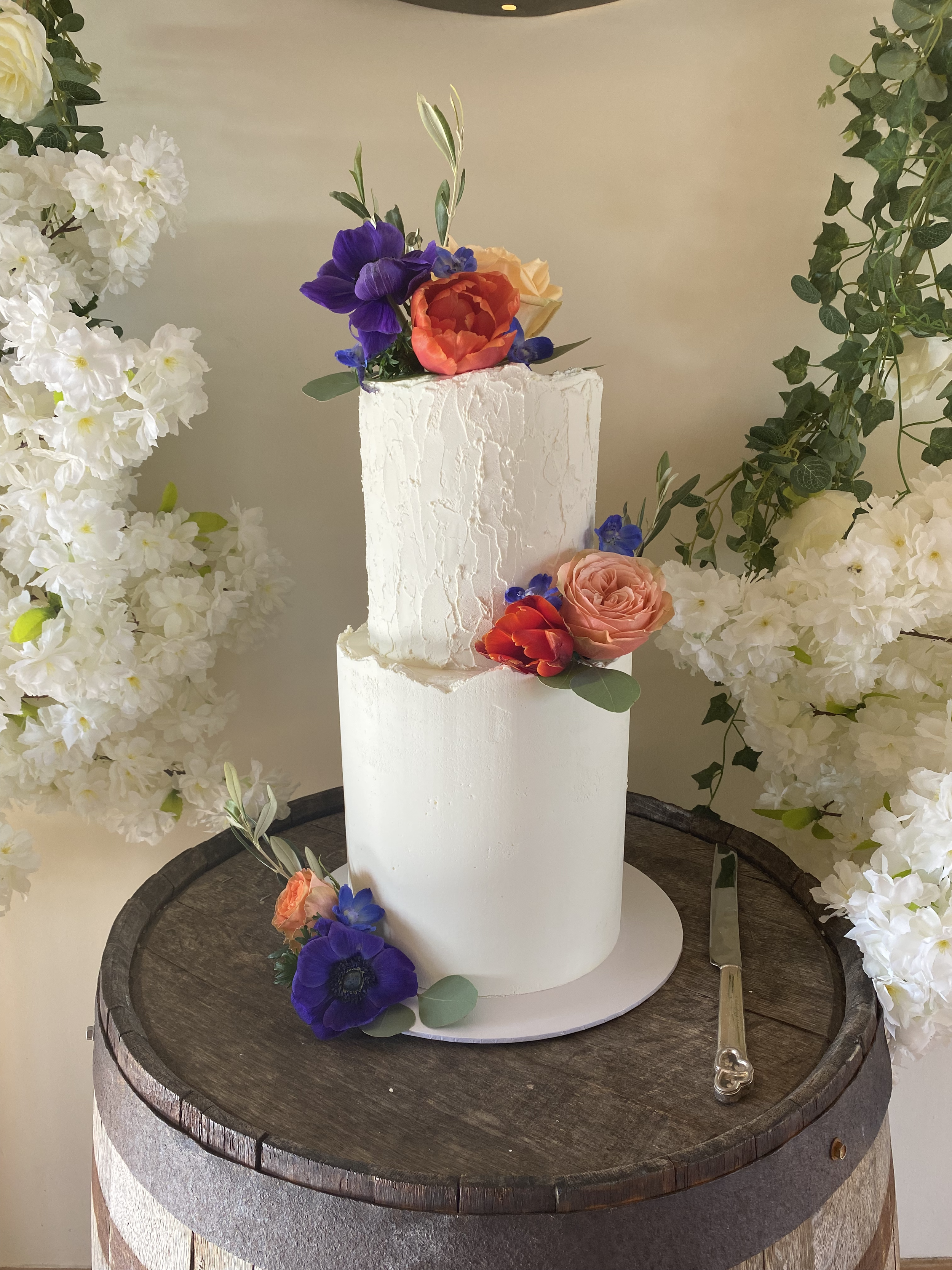 A white textured wedding cake decorated with purple, orange, peach, and cream-colored flowers, surrounded by white hydrangeas and green leaves on a wooden table.