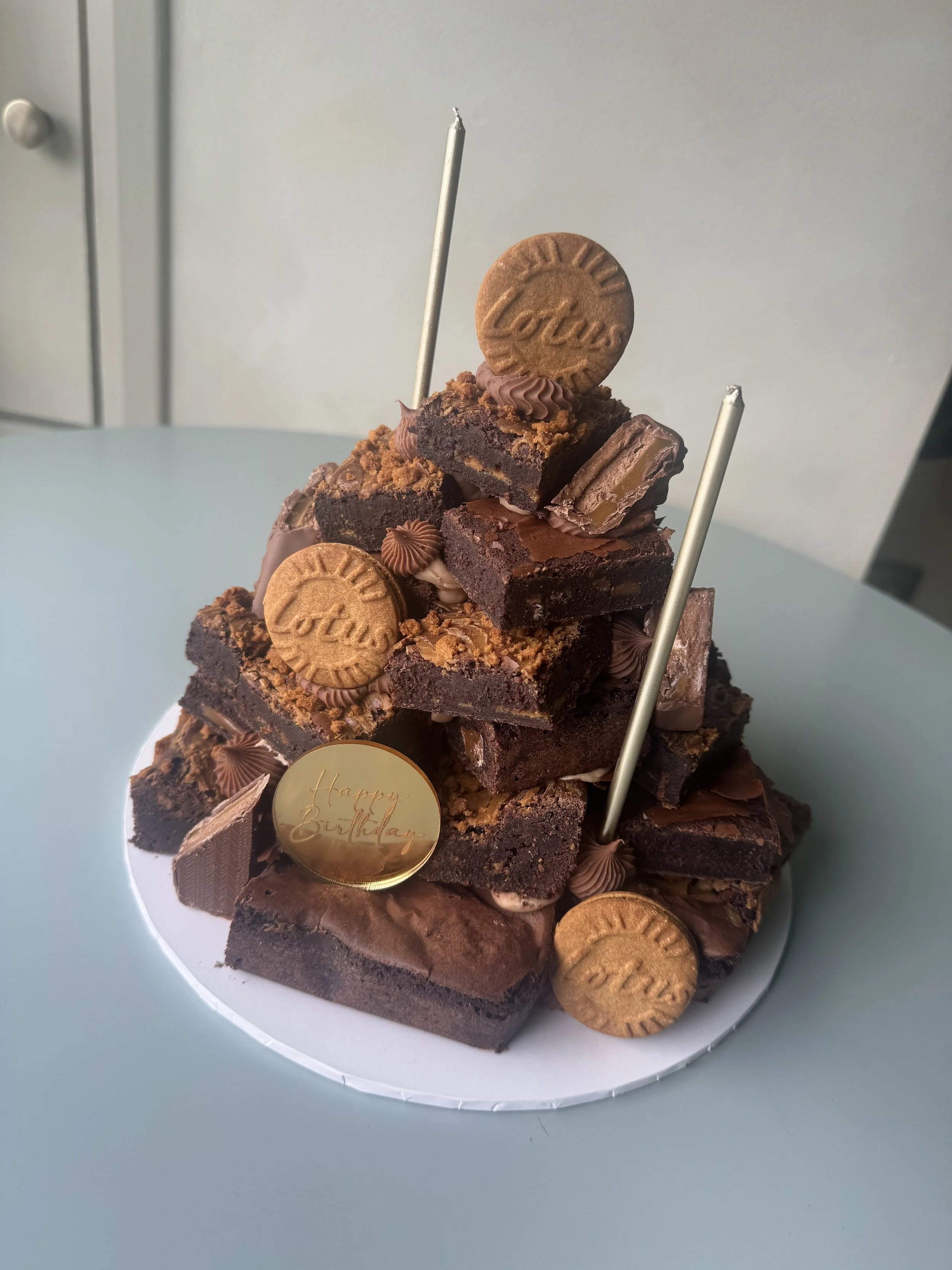 A multi-layer chocolate birthday cake decorated with chocolate squares, Lotus cookies, and gold accents.