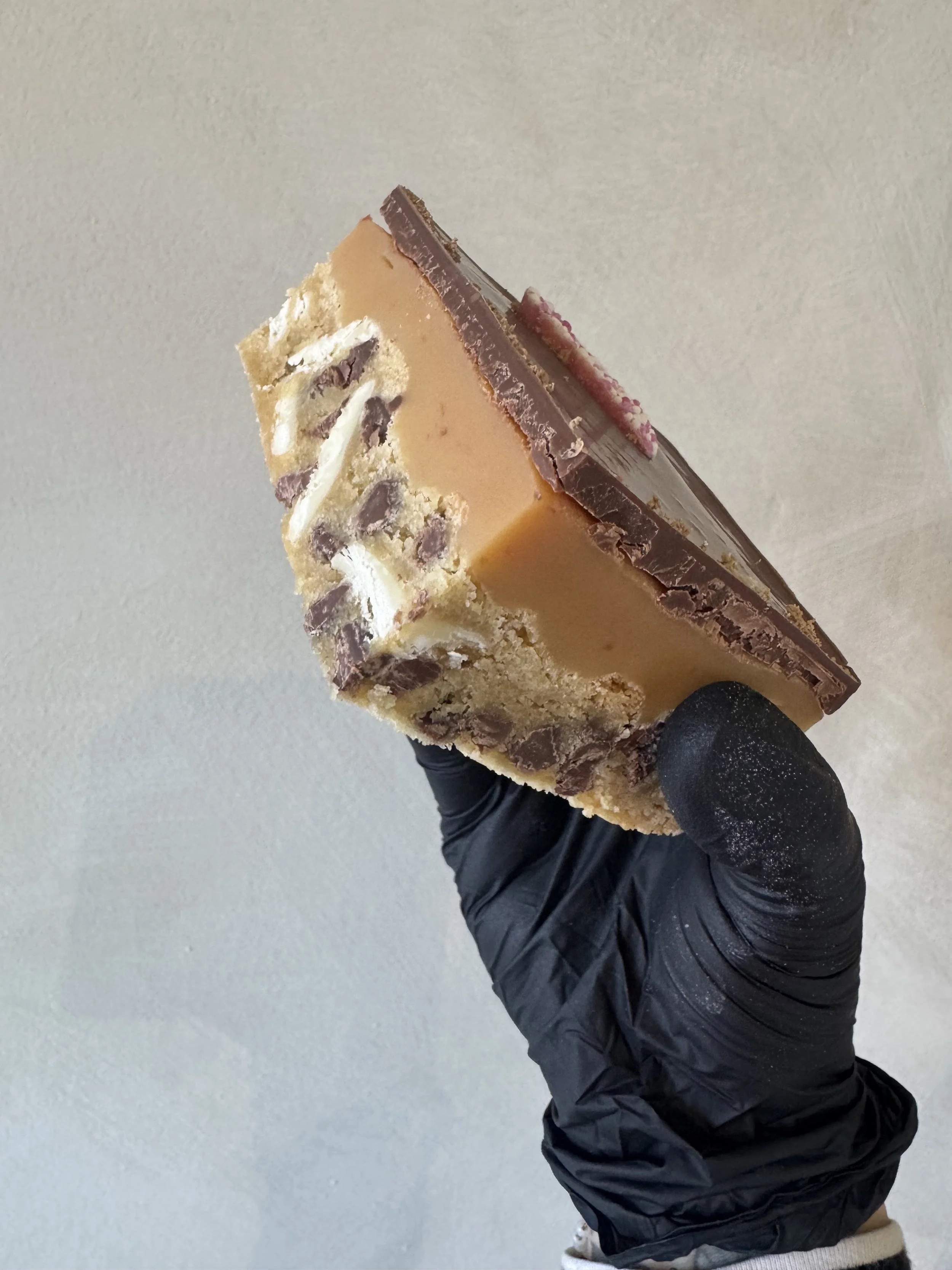A person wearing a black glove holding a slice of caramel and chocolate layered dessert with nuts and chocolate chips on top against a plain background.