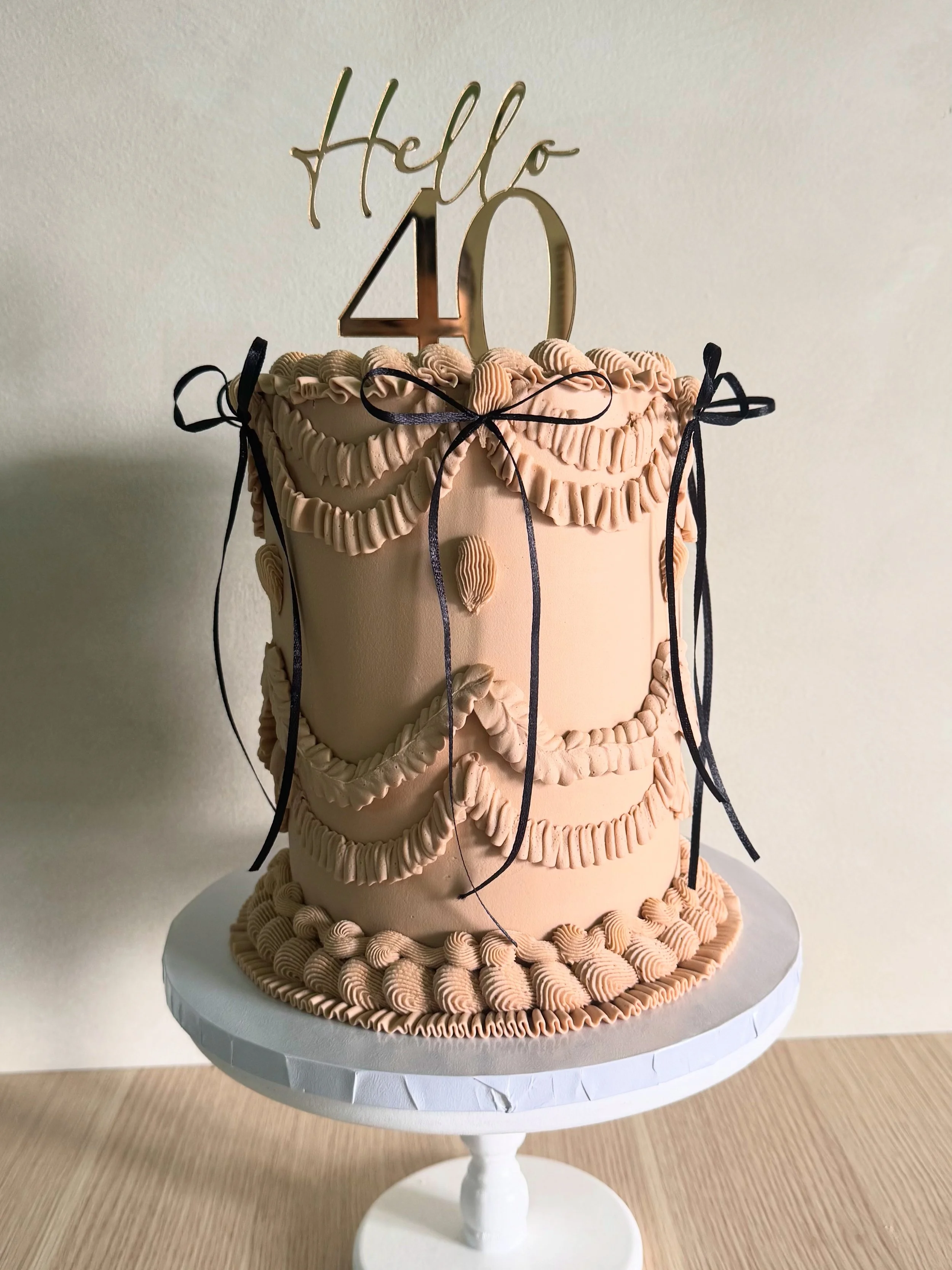 A tall, peach-colored cake decorated with piped rosettes and ribbons, topped with a gold 'Hello 40' cake topper, placed on a white cake stand.