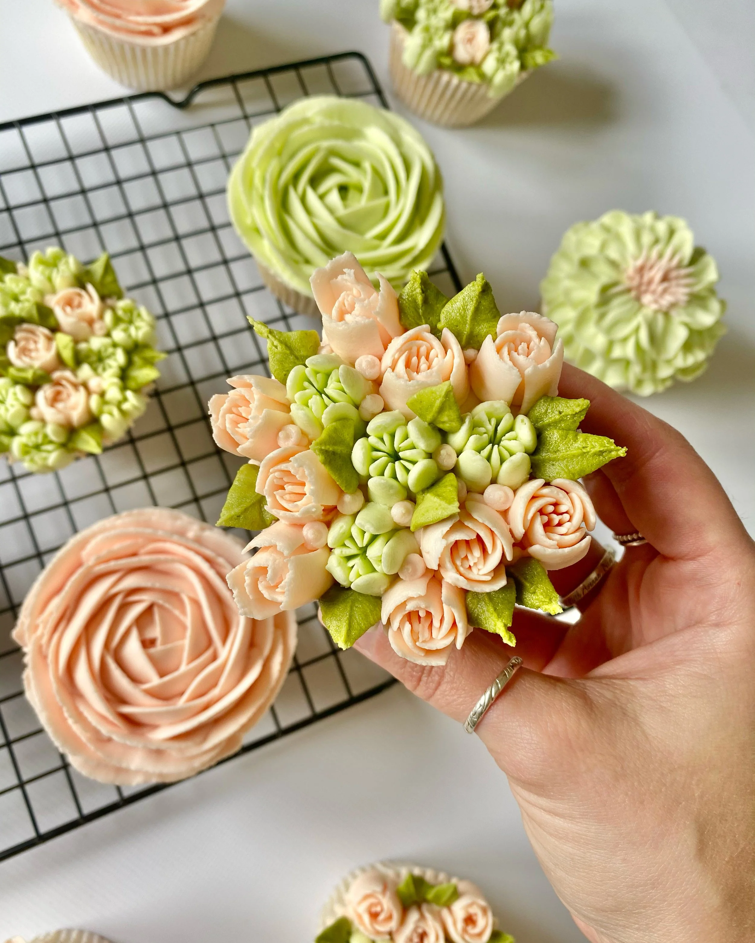 Hand holding a cupcake decorated with pastel pink roses, green leaves, and small pale green buds, with other decorated cupcakes and a cooling rack in the background.