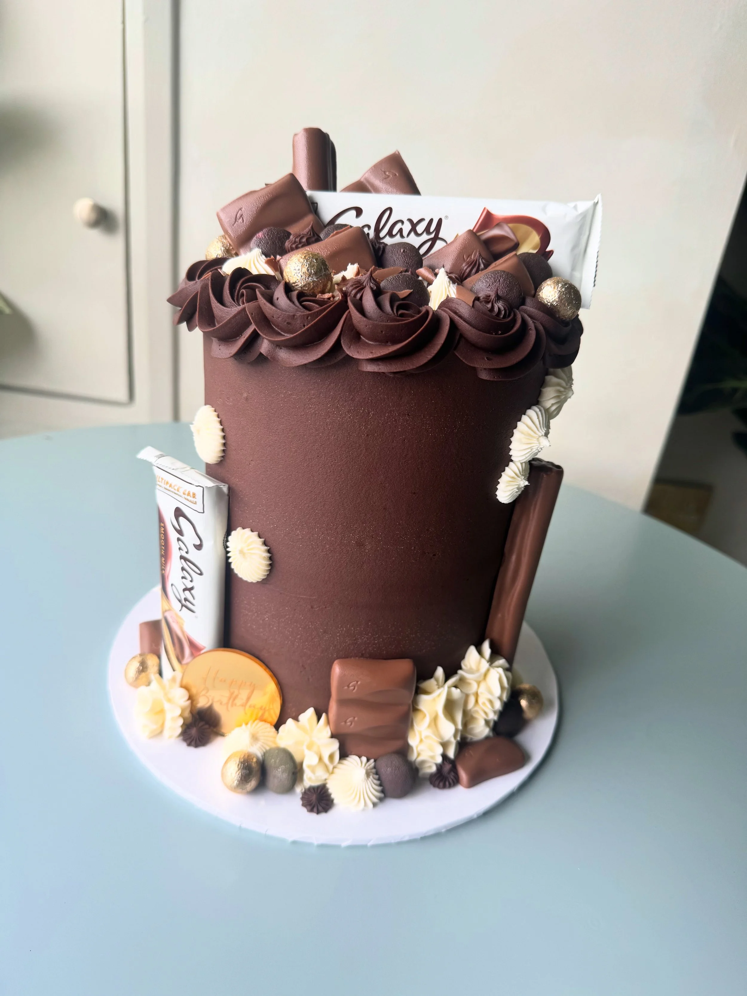 A tall chocolate birthday cake decorated with chocolate and vanilla icing, chocolates, gold foil-wrapped candies, and a Galaxy chocolate bar on top, surrounded by piped icing roses and other confections.