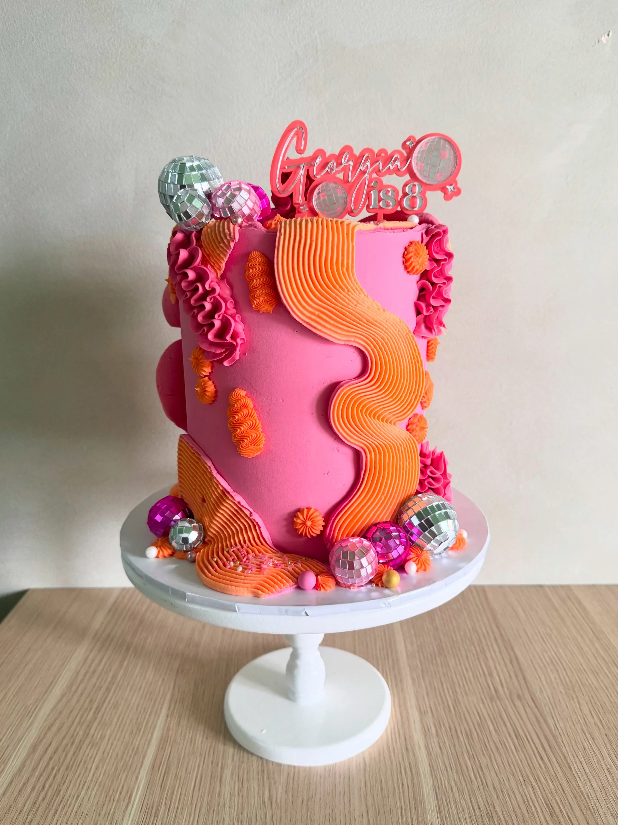 Pink birthday cake with orange and pink frosting decorations, topped with a pink 'Gorgina is 8' cake topper, surrounded by disco ball ornaments, placed on a white cake stand on a wooden surface.