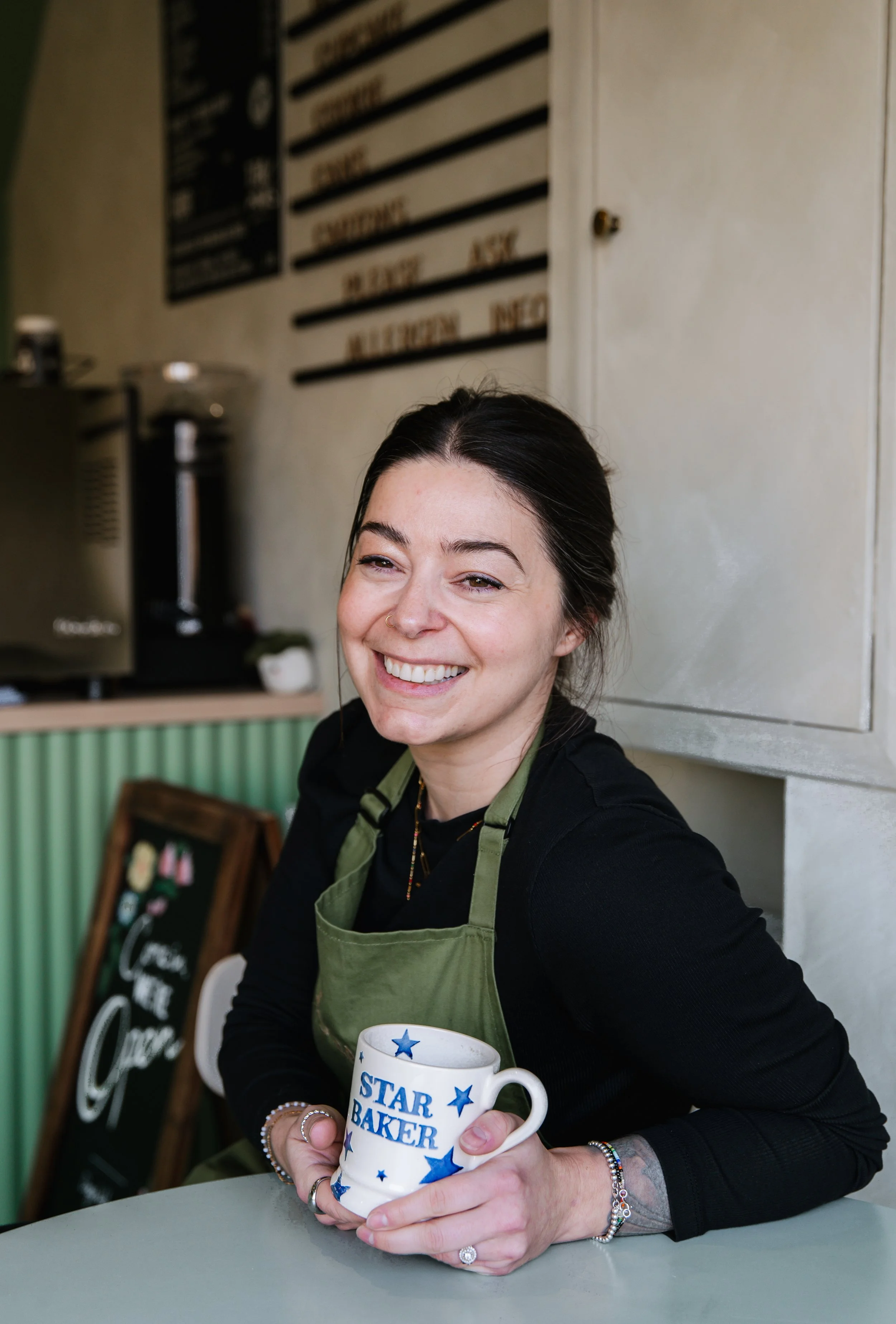 A smiling woman with dark hair tied back, wearing a black shirt and a green apron, holding a white mug with blue stars and the text 'STAR BAKER' in a cafe or bakery setting.