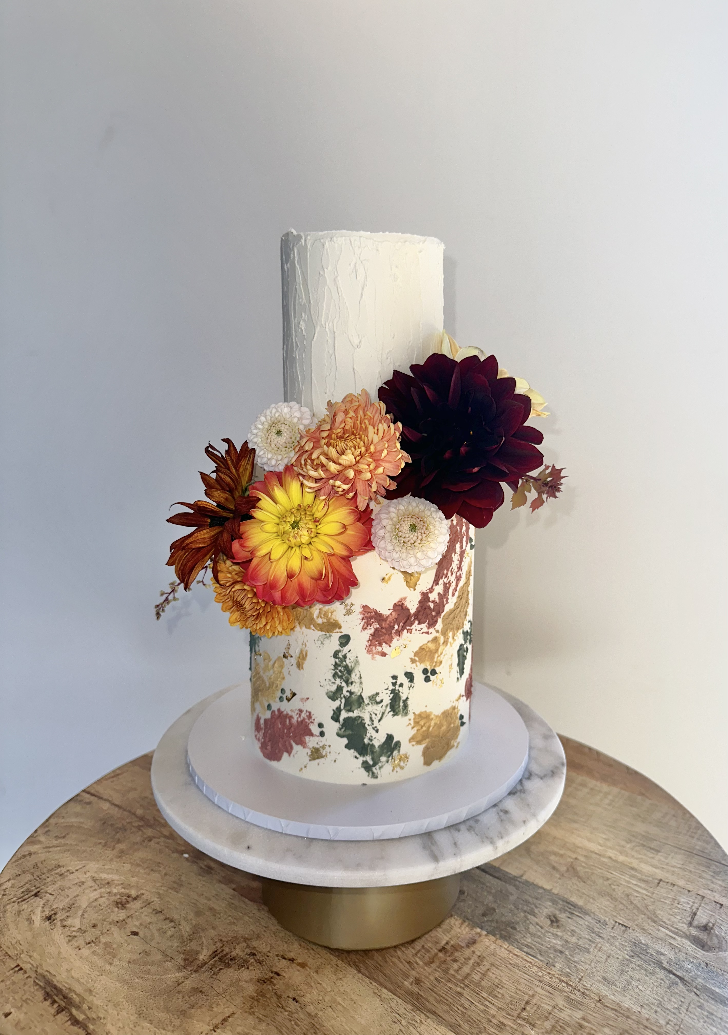 A tall white cake decorated with colorful flowers including yellow, orange, and dark red blooms, sits on a white cake stand on a wooden surface. The cake has an artistic, watercolor-like design on its side.