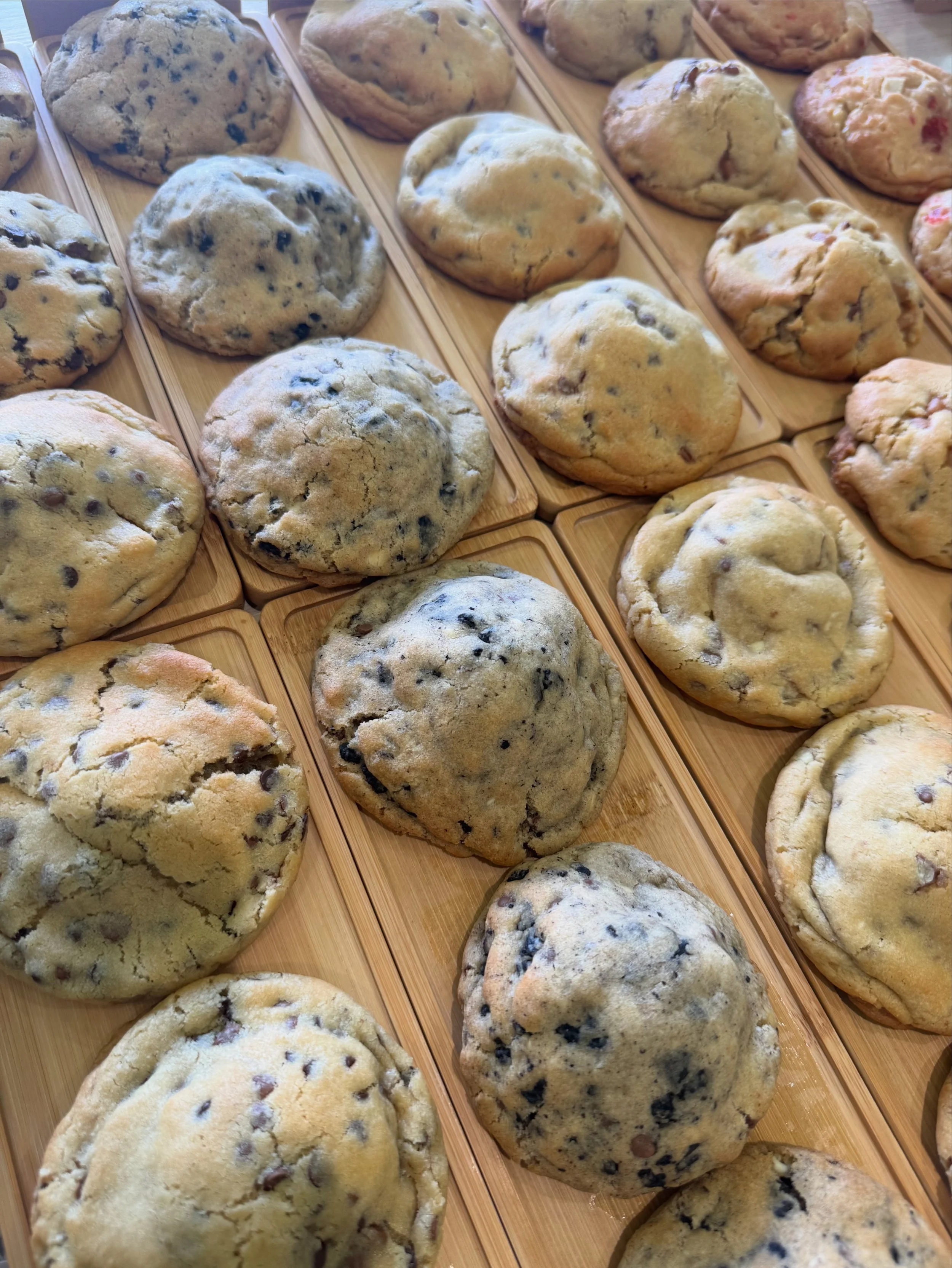 Multiple freshly baked cookies on wooden boards, including chocolate chip, mixed berries, and chocolate varieties.