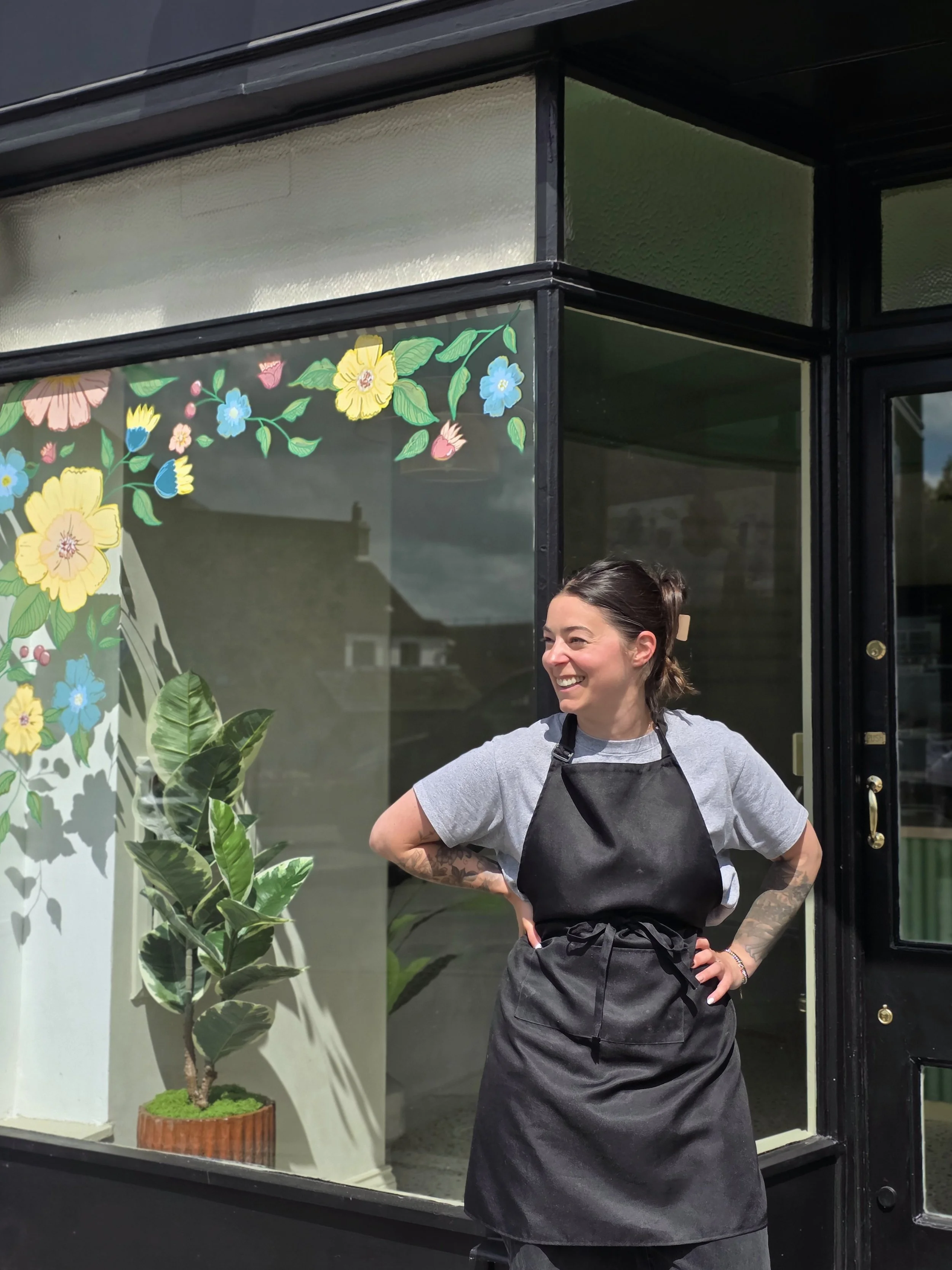 Smiling woman in a gray t-shirt and black apron standing outside a storefront with a glass window featuring a floral design and a potted plant nearby.