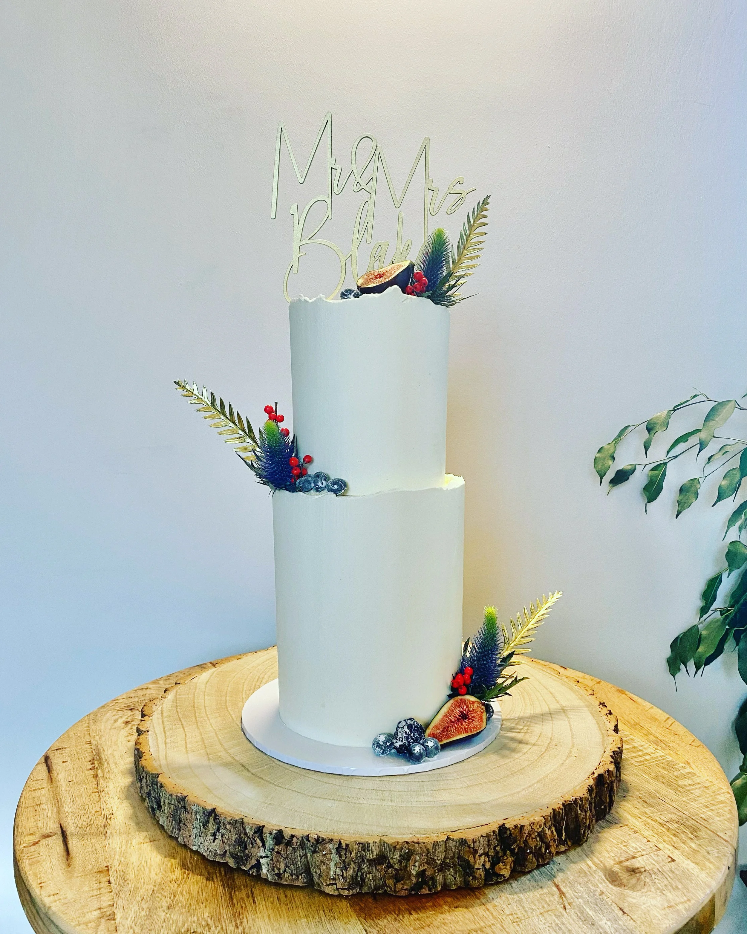 A two-tier white cake with fresh fruit and greenery decorations, topped with a gold "Mr & Mrs Black" cake topper, displayed on a wooden tree slice.