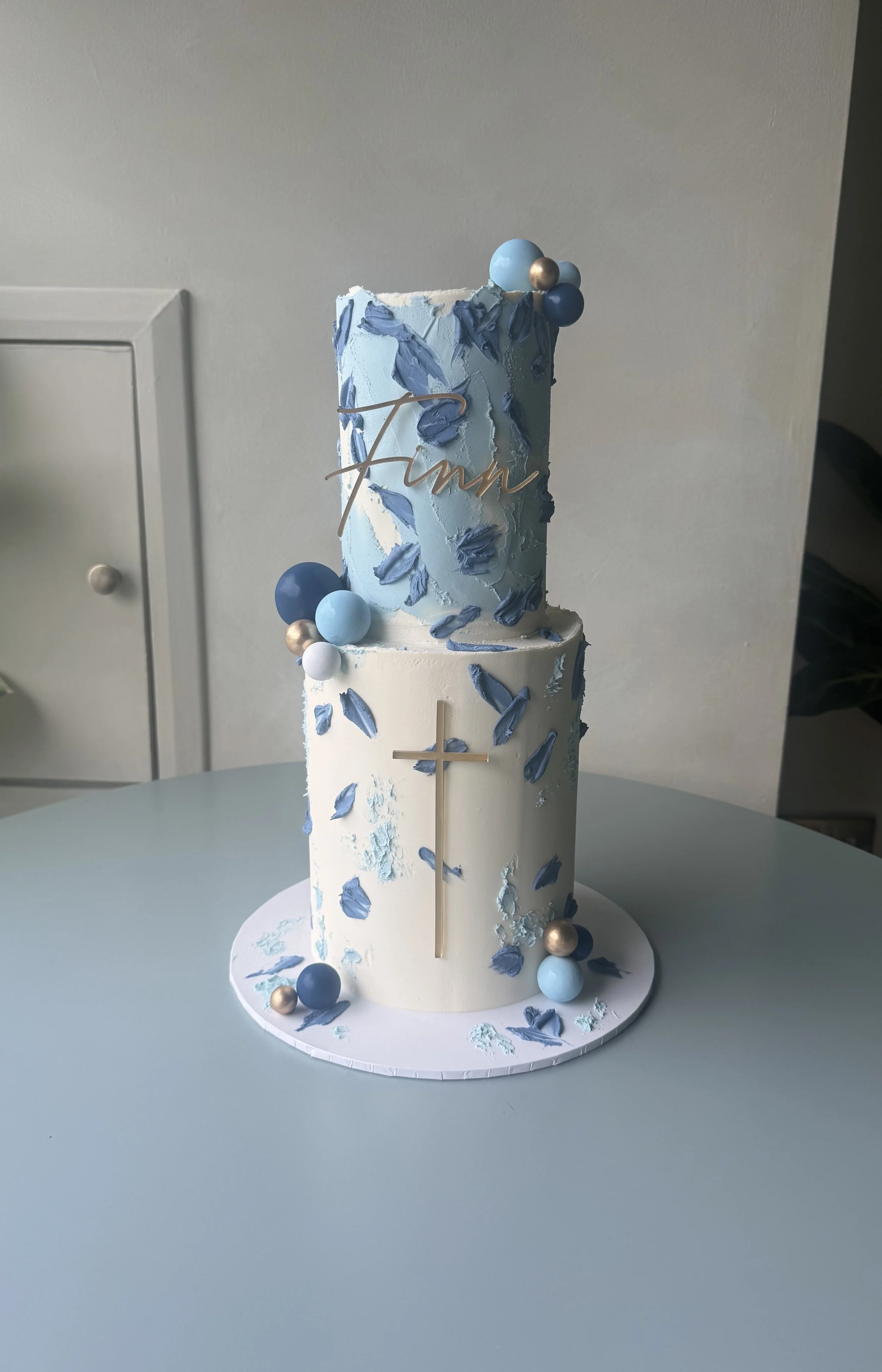 A tall, multi-tiered cake decorated in shades of blue, white, and gold, with abstract blue icing patterns, spheres, and a gold cross, set on a white base with scattered blue and gold accents.