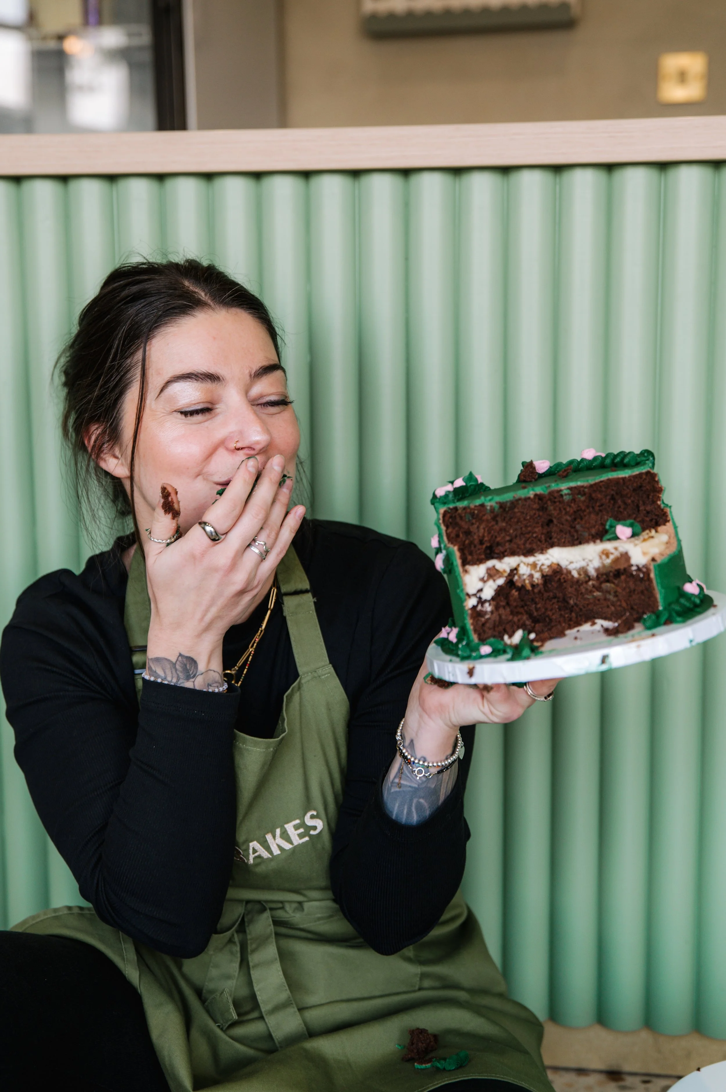 A woman with dark hair, wearing an apron, is holding a slice of chocolate cake with green icing and pink decorations, and appears to be about to eat it.