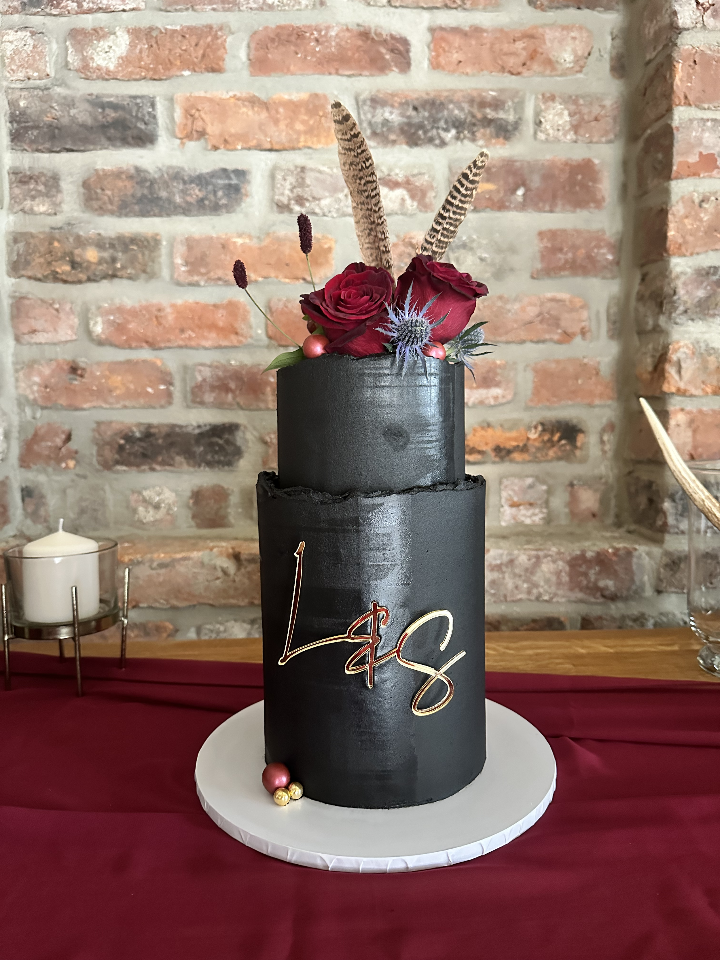 Black cylindrical cake with a gold handwritten 'LC' on the side, decorated with red roses, purple thistle, and feathers, placed on a white round cake board on a red tablecloth with a brick wall background.