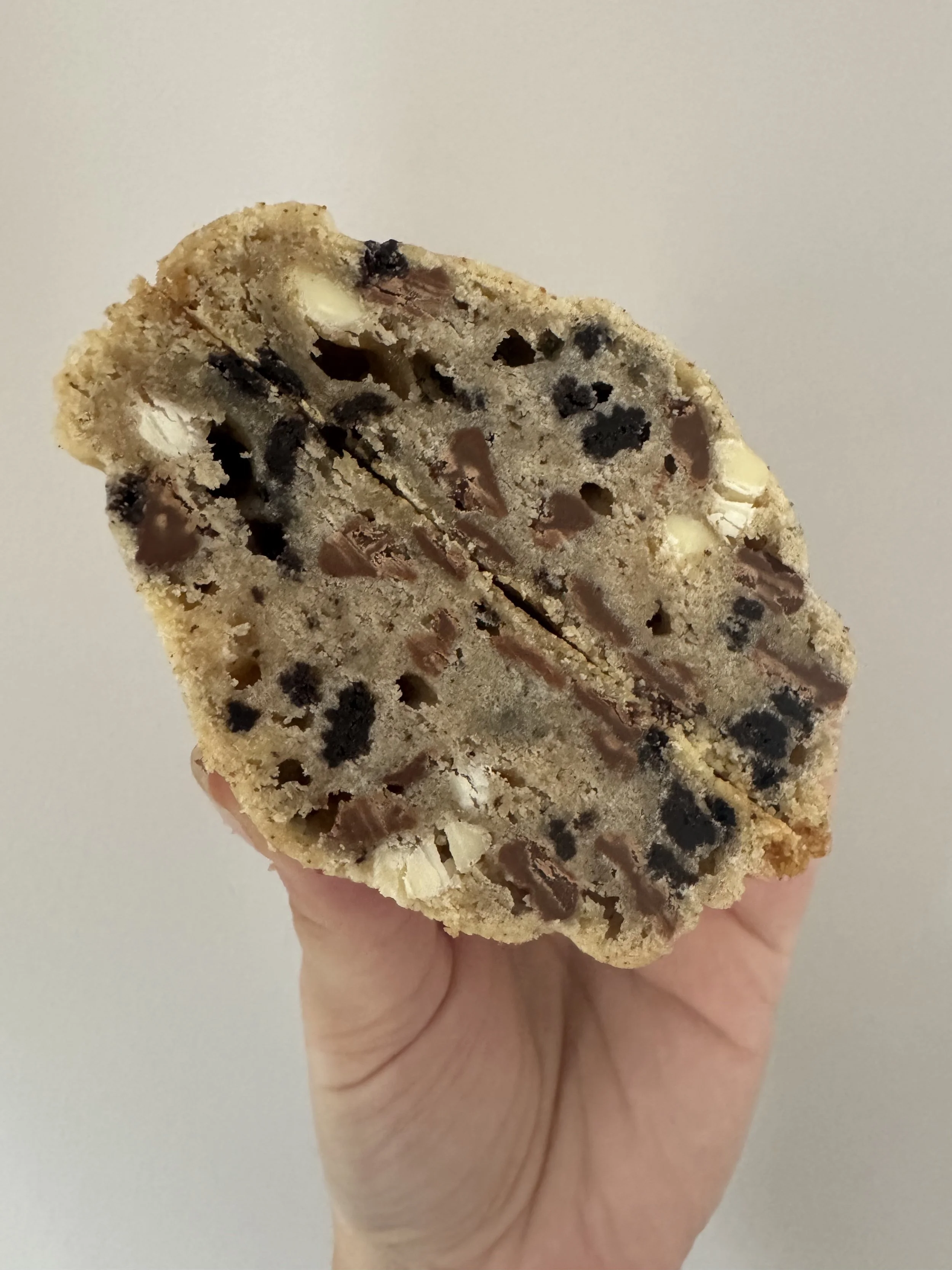A hand holding a cookie with chocolate chips and white chocolate chunks against a plain light background.
