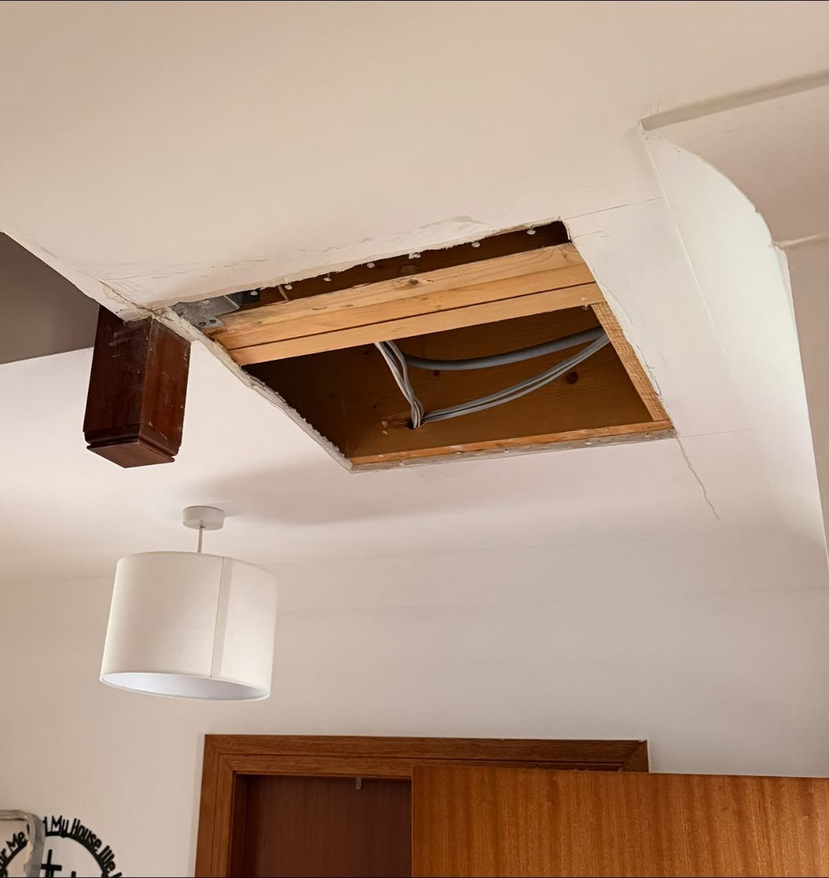 Ceiling with an opening revealing electrical wires and wooden support beams, with a hanging light fixture and a wooden door.