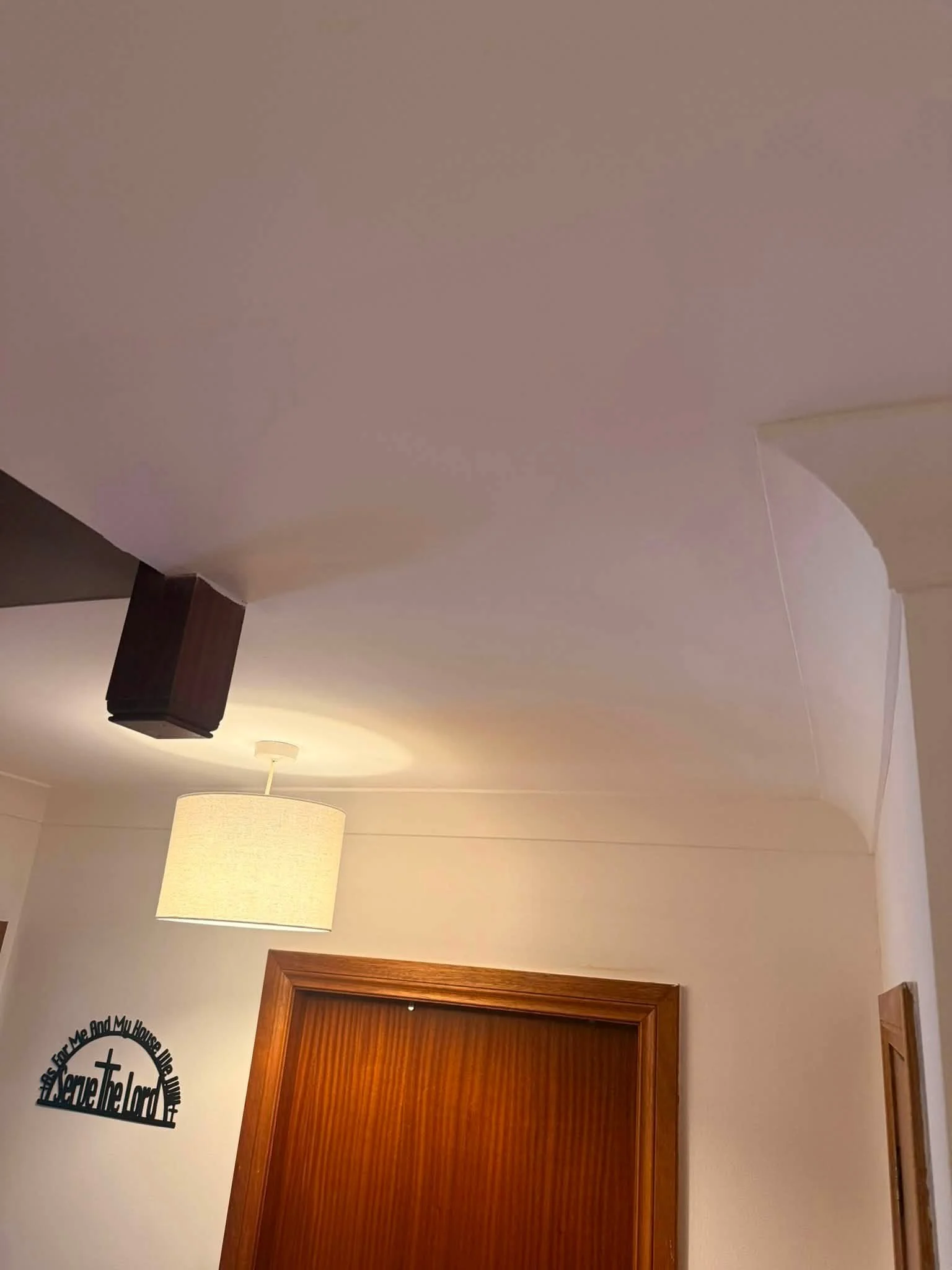 Image of a ceiling with a dark wooden beam, a hanging light fixture with a beige lampshade, a wall decoration with text and a cross, and a door with a wooden frame.